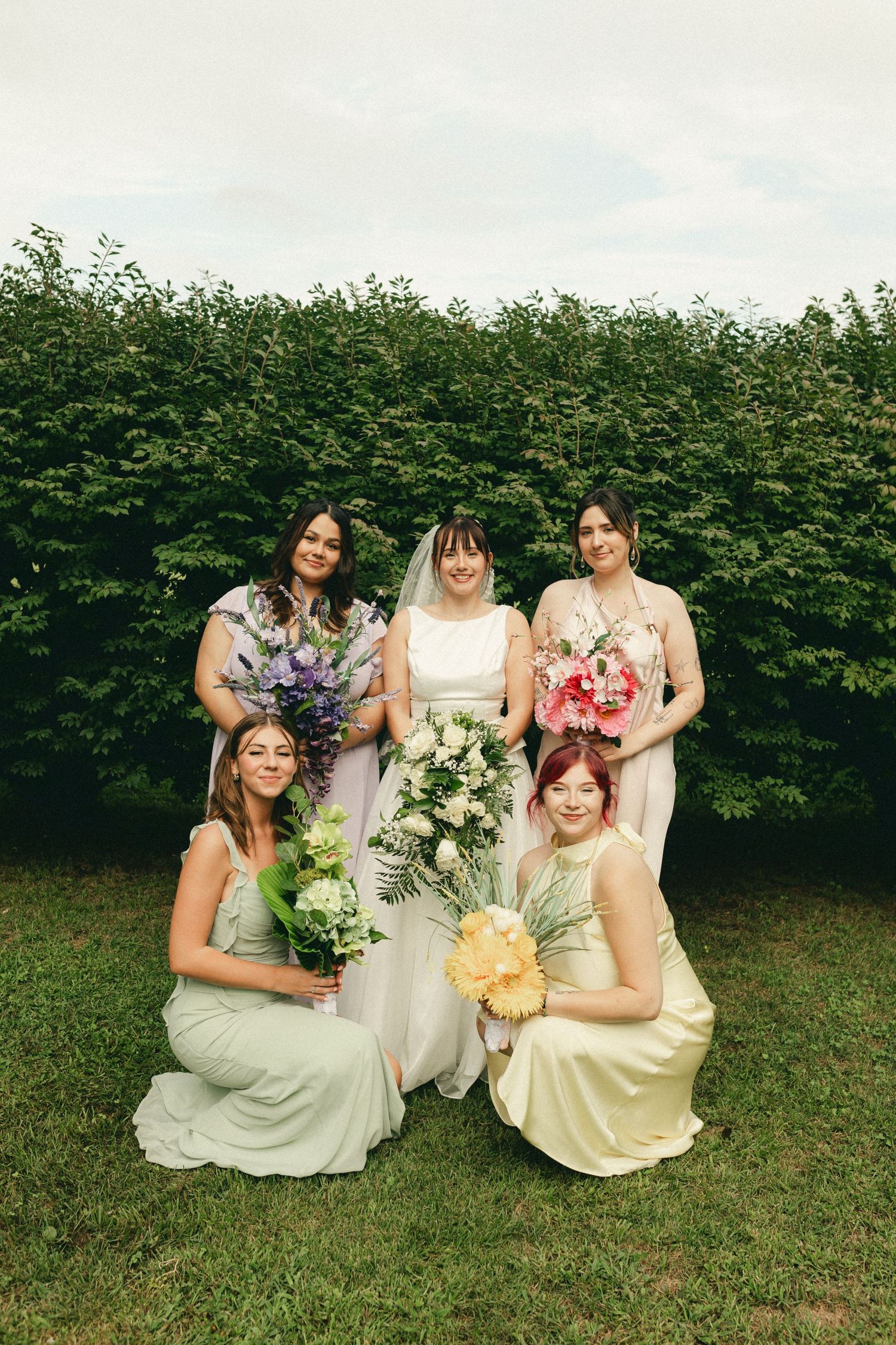 Bride in white gown with bridesmaids in pastel dresses holding colorful bouquets outdoors.