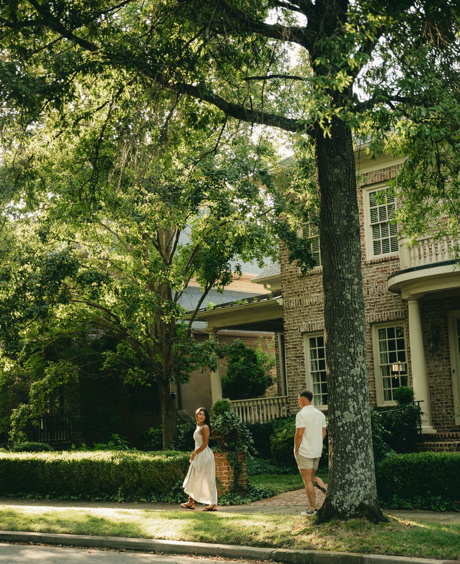 Couple standing apart near large tree outside brick home during engagement session.