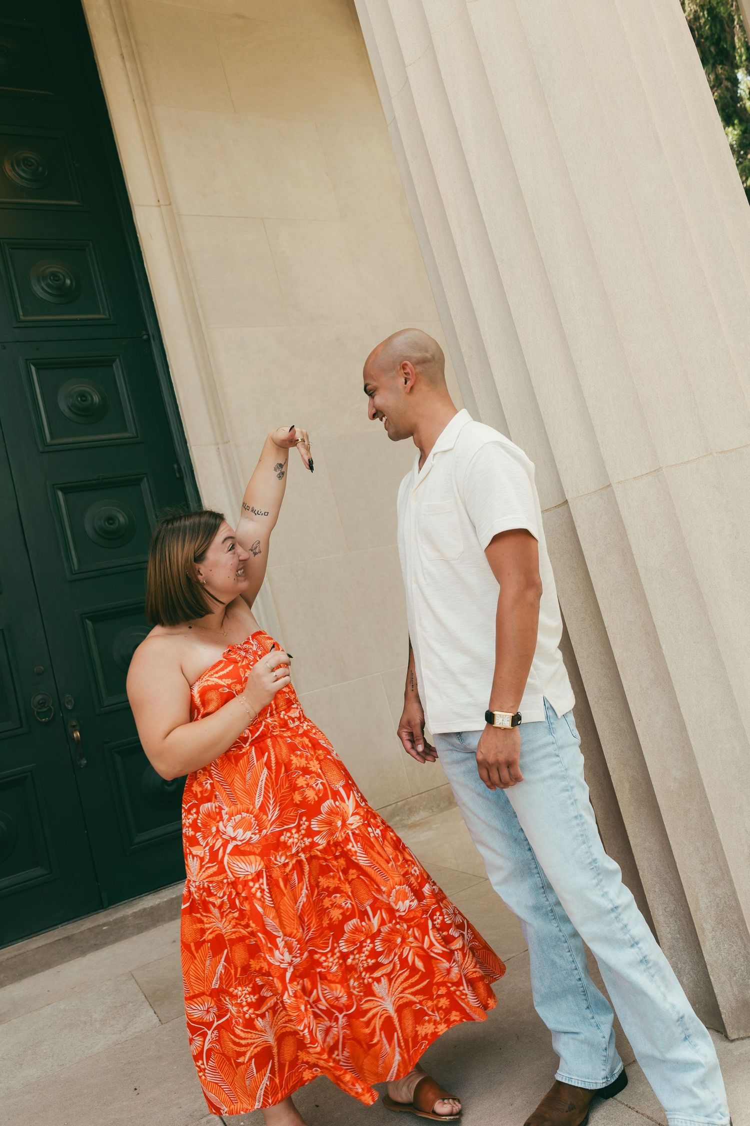Couple laughing and spinning together outdoors near stone pillars, woman wearing a vibrant orange floral dress.