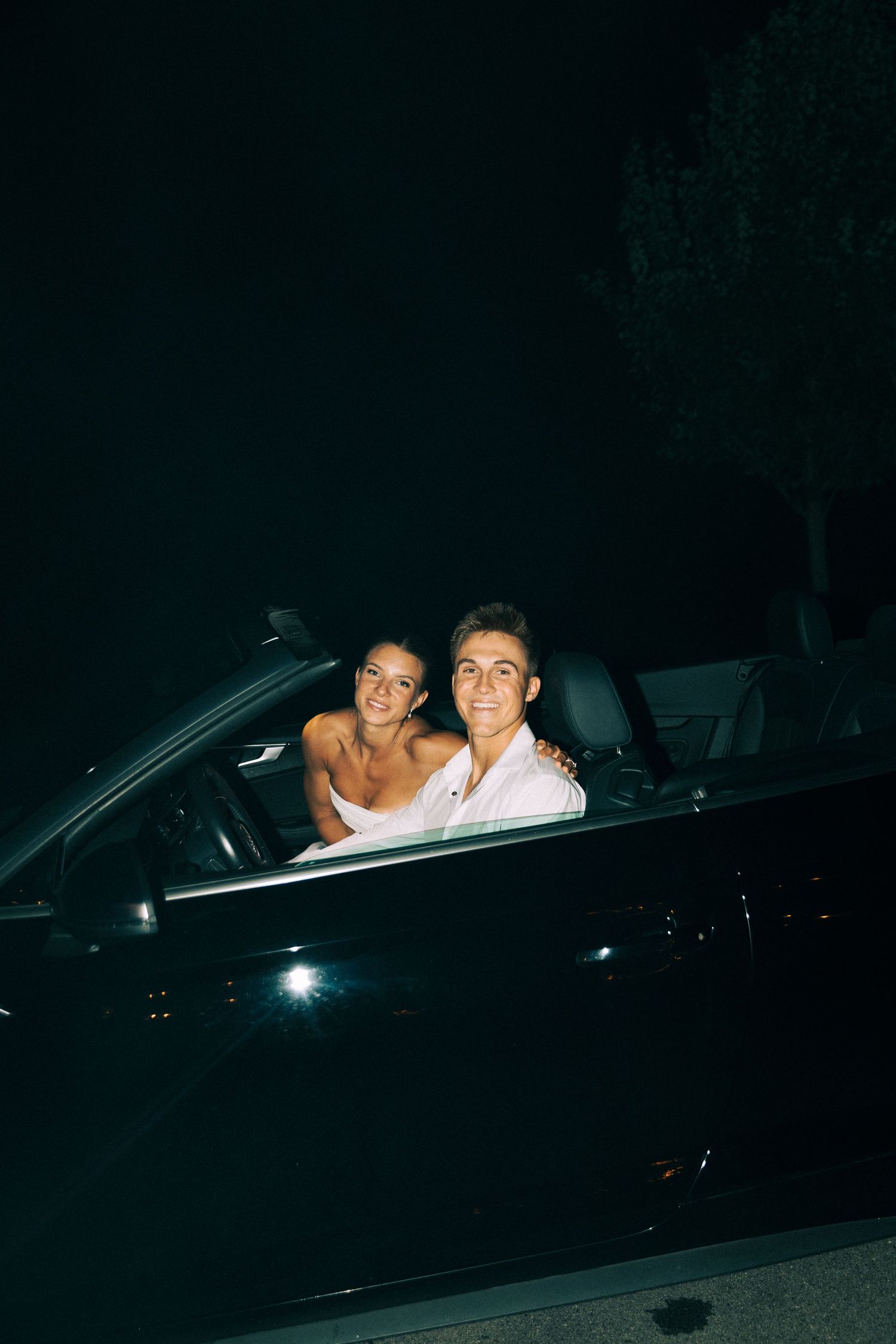 Newlywed couple smiles together in a convertible car at night after their wedding reception.
