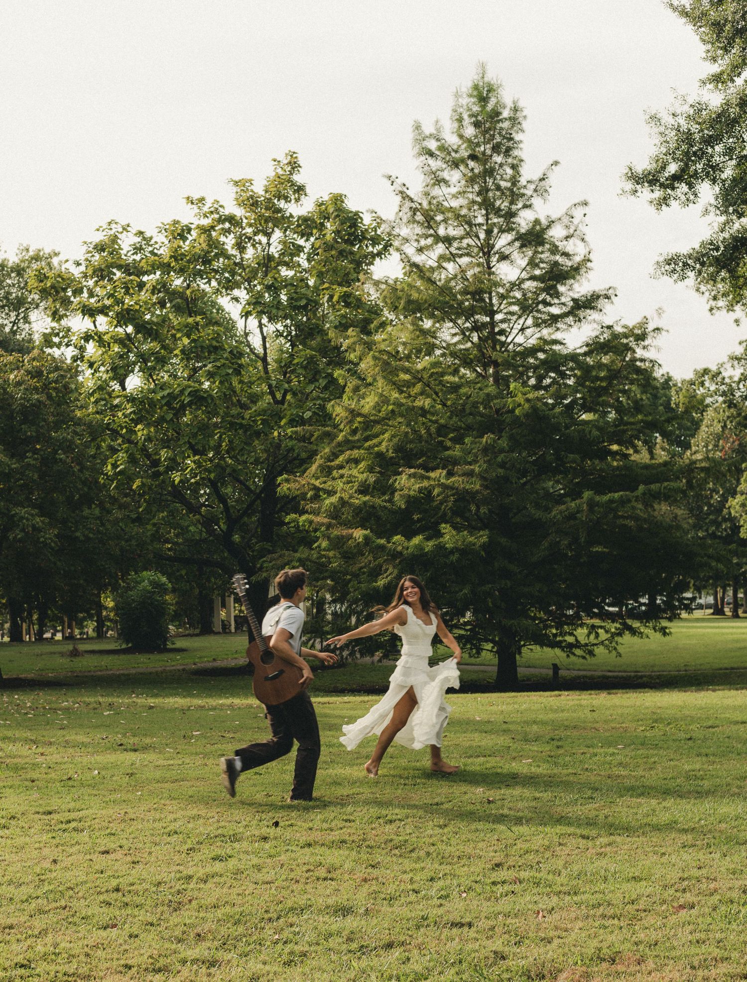 Bride and groom joyfully running hand-in-hand across a lush green park surrounded by tall trees on their wedding day.