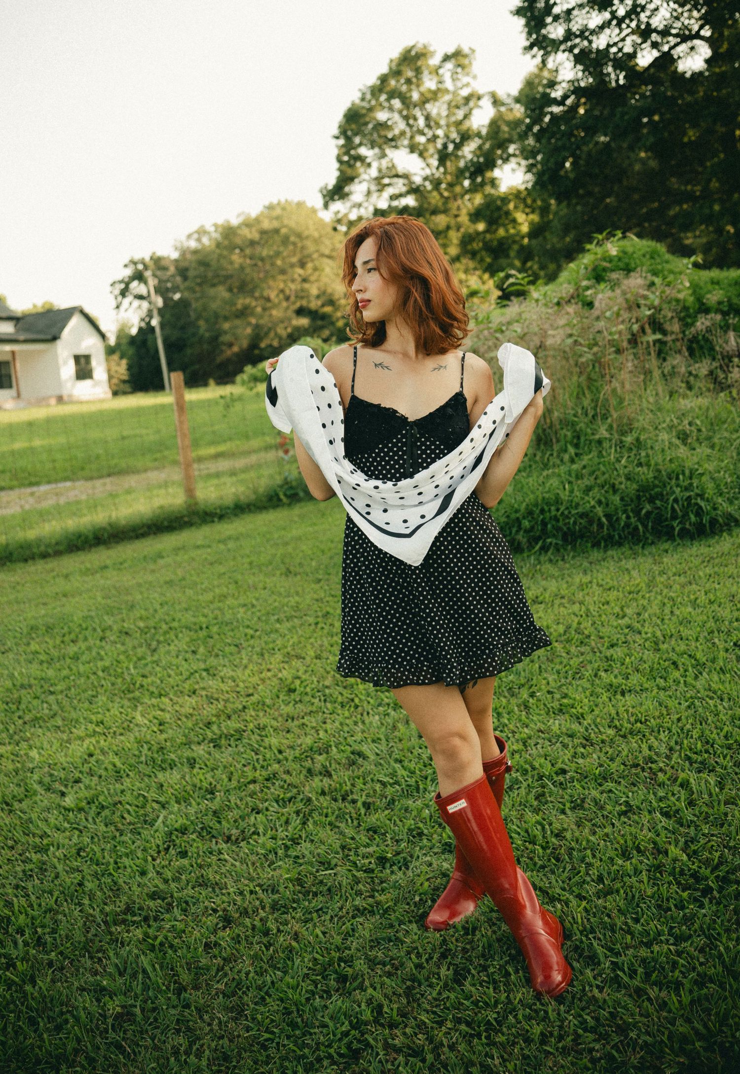 Young woman in polka dot dress and red Hunter boots posing in green field.