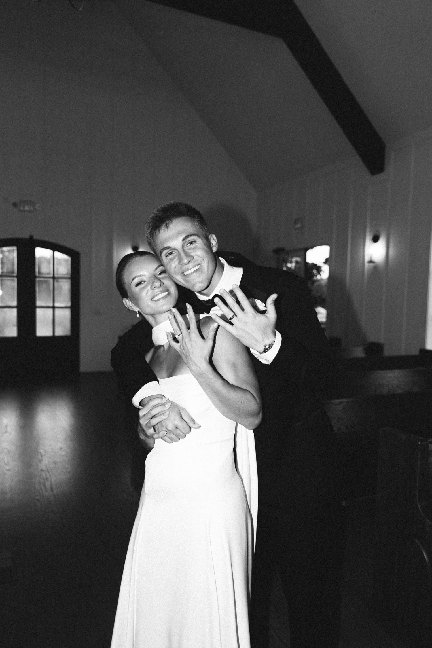 Newlyweds embrace and show off wedding rings in black and white reception photo inside elegant venue.