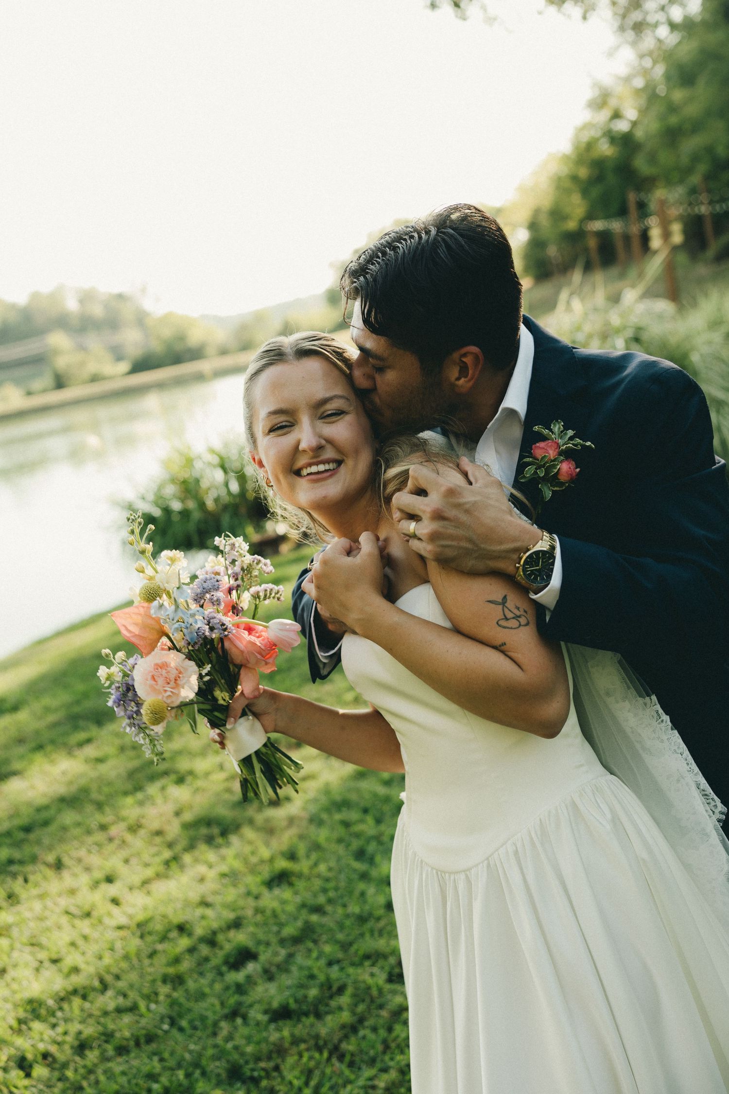 Groom in navy suit kissing bride's cheek as she laughs holding colorful bouquet by a river.