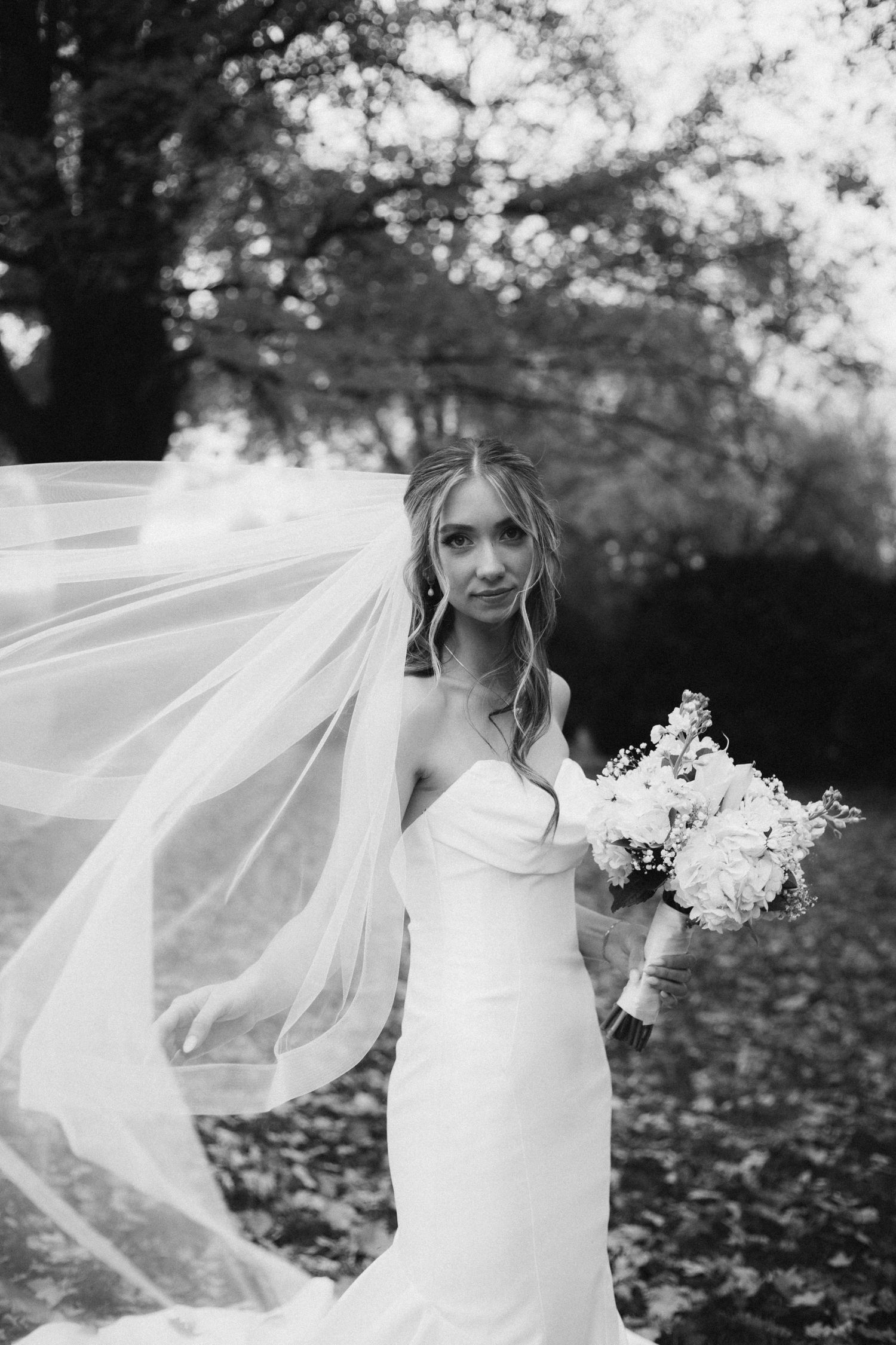 Black and white bridal portrait with veil flowing in wind, bride holding bouquet in strapless gown outdoors.