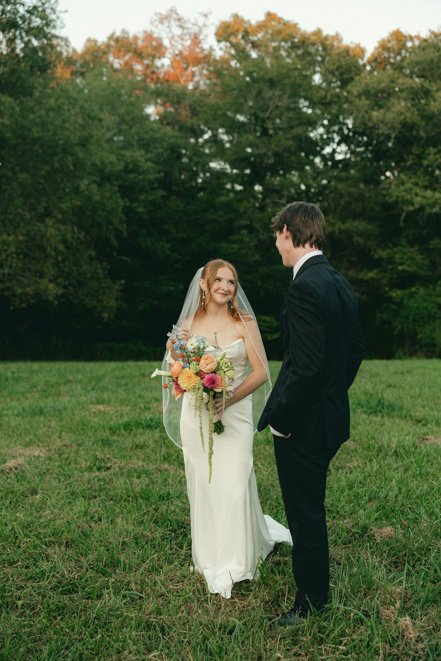 Bride in white strapless gown with veil holds colorful bouquet facing groom in black suit on a green lawn.