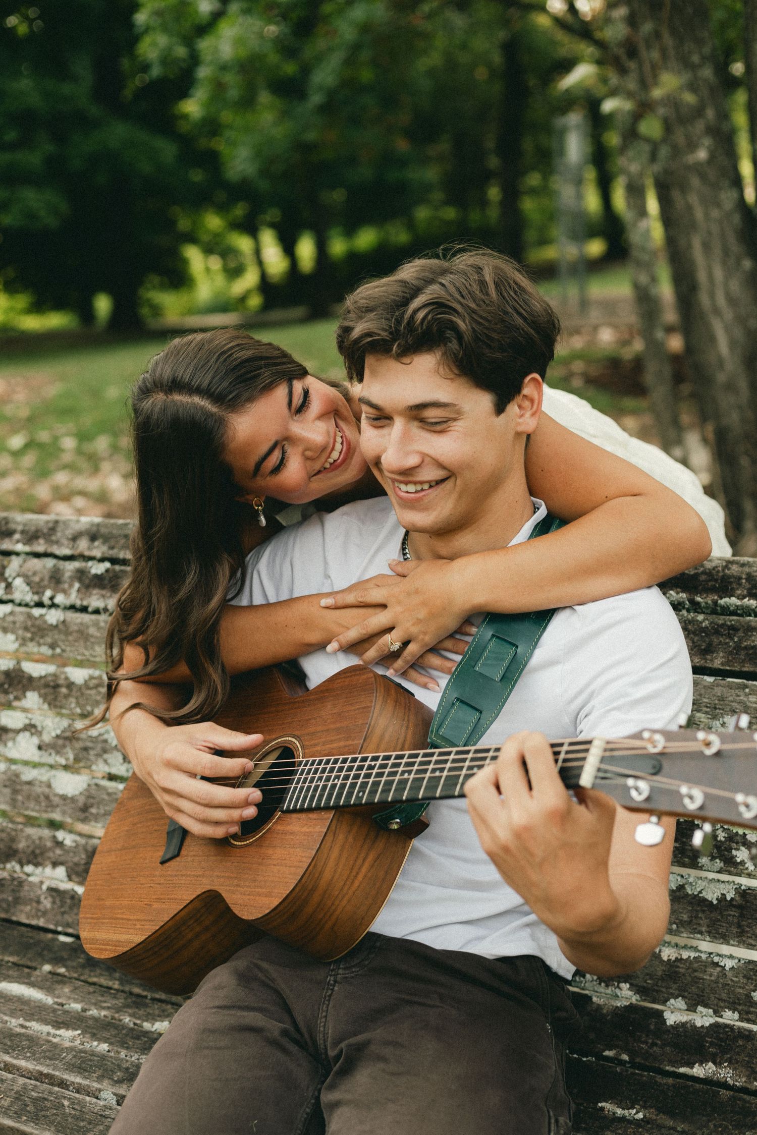 Happy couple on a park bench, woman hugging man from behind as he plays acoustic guitar.