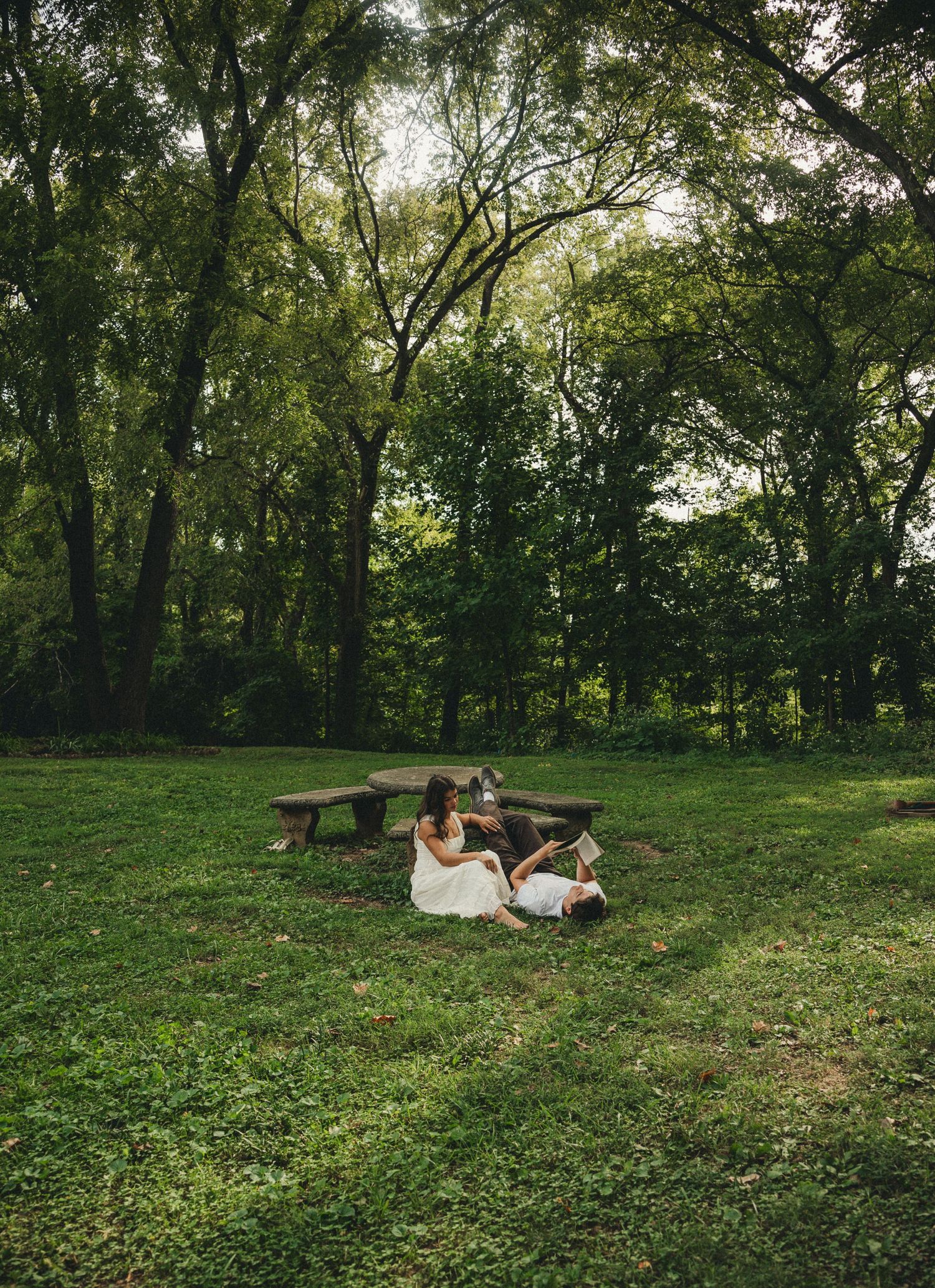 Two people in white dresses sit on lush green grass near a stone picnic table surrounded by tall trees in a serene park.