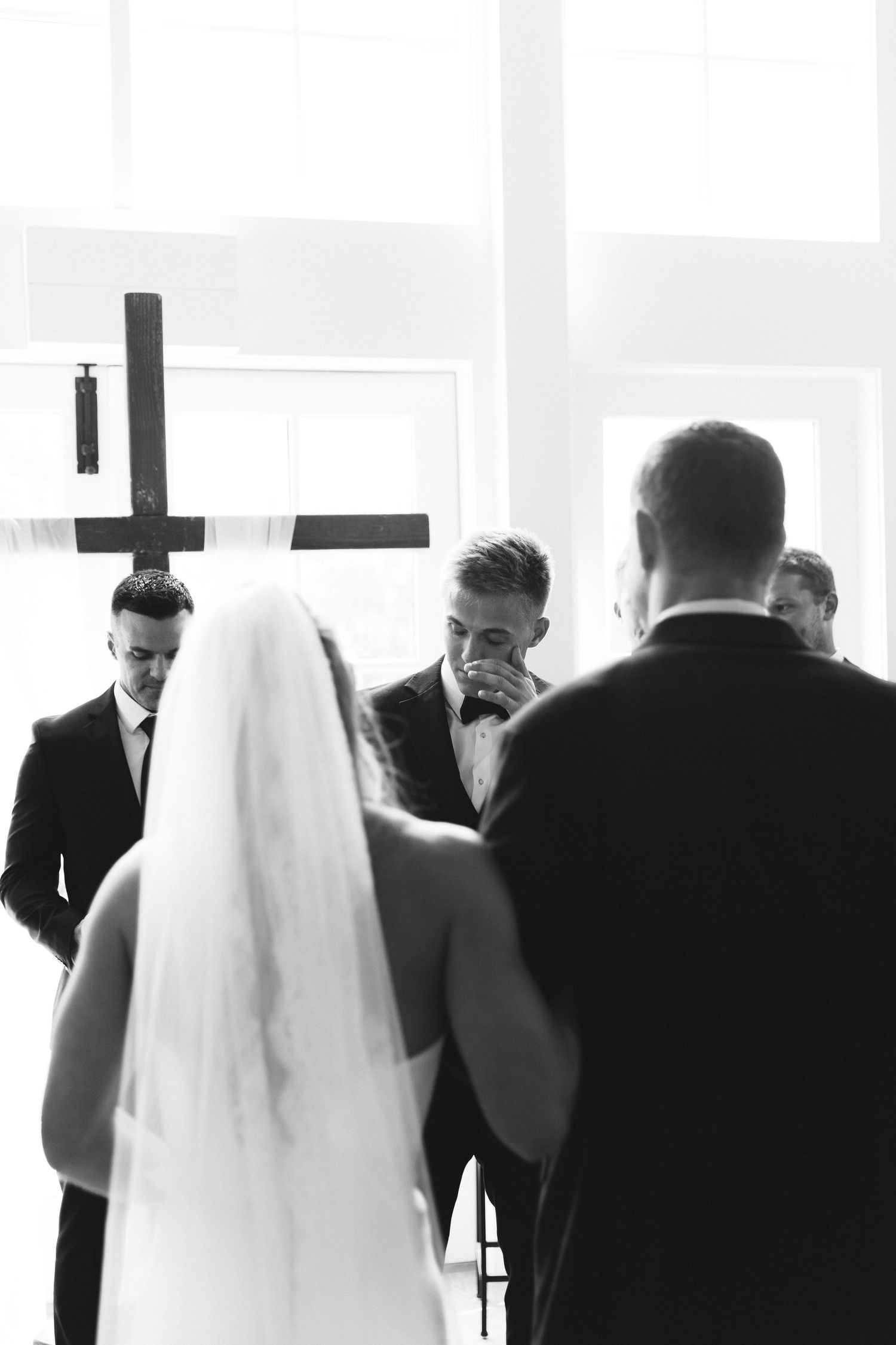 Black and white photo of bride and groom at wedding altar with cross and emotional groomsmen.