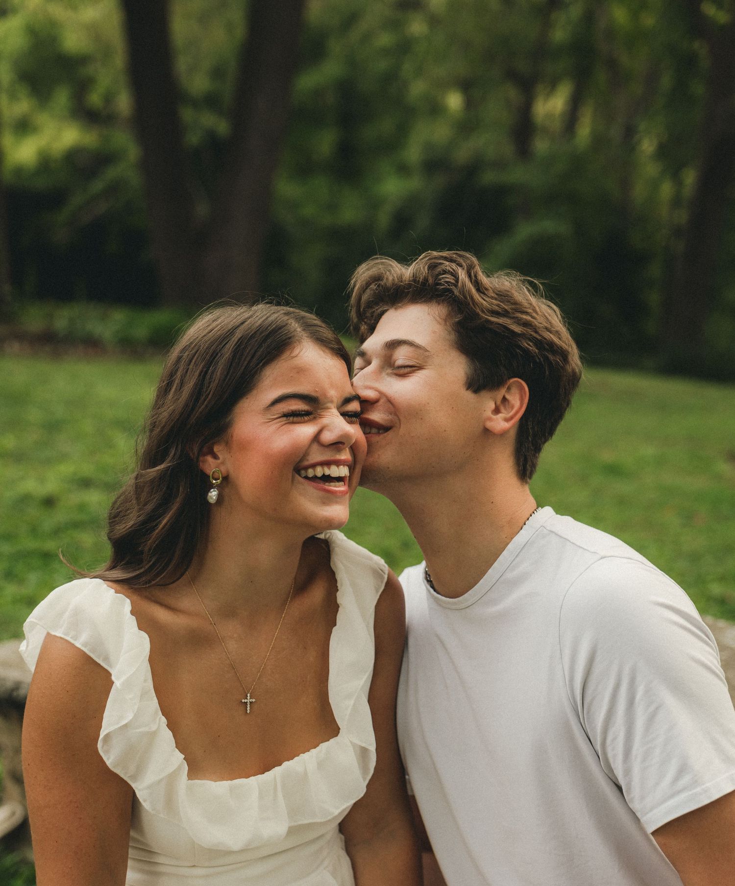 Joyful couple sharing a kiss on the cheek outdoors, woman laughing in white ruffled dress.