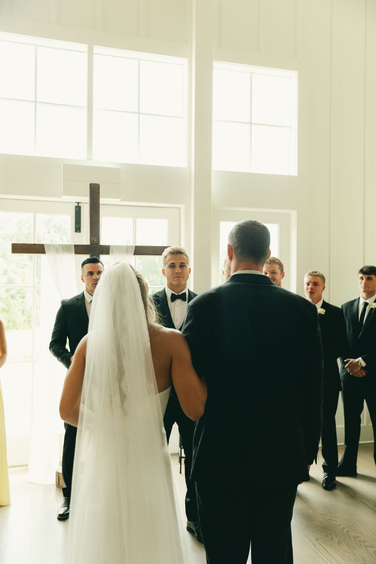 Bride in white gown and veil walks down aisle toward groom at altar with wooden cross in bright chapel.