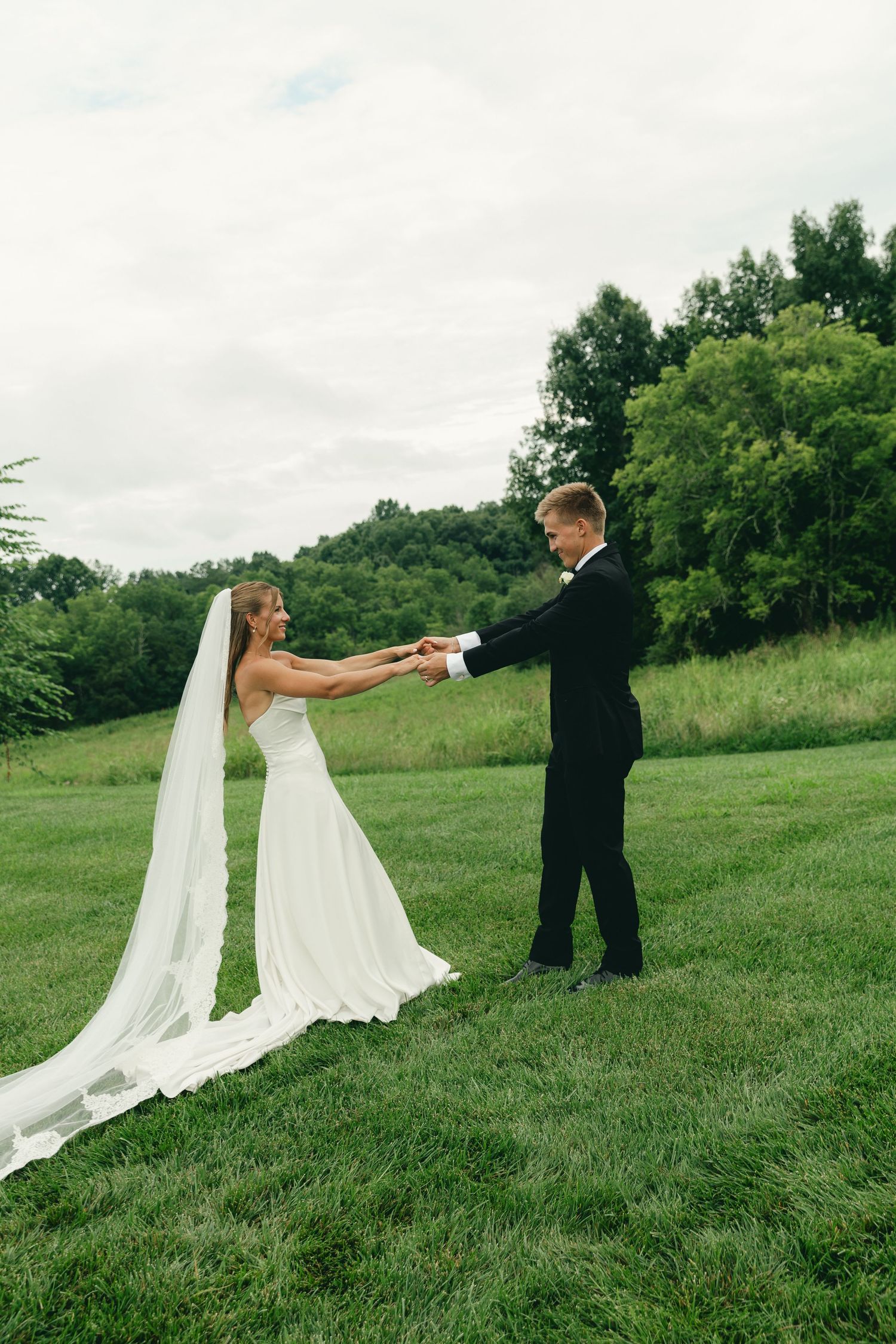 Bride and groom hold hands on a green lawn, her long white veil trailing behind with rolling hills in the background.