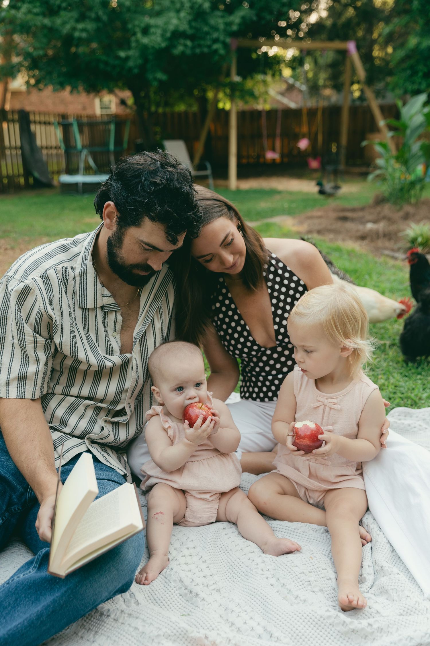 Family reading book together on blanket outdoors with two babies eating apples in backyard.