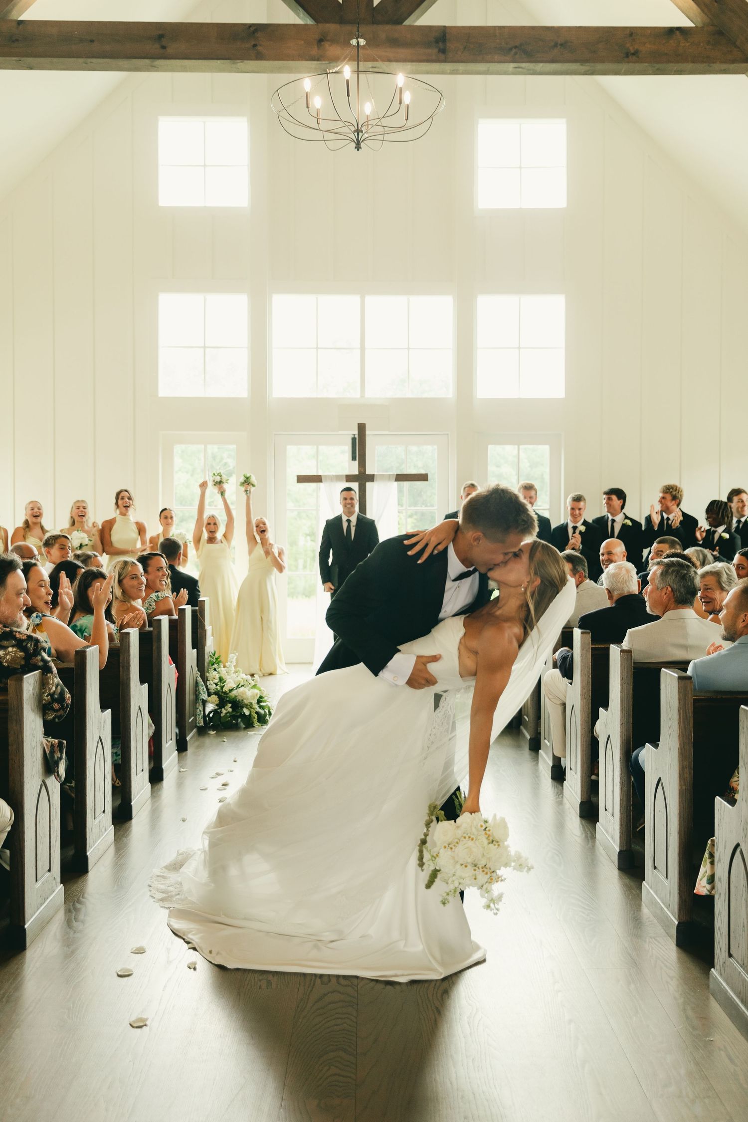 Groom dips bride for romantic kiss at altar after wedding ceremony in white chapel with wooden cross.