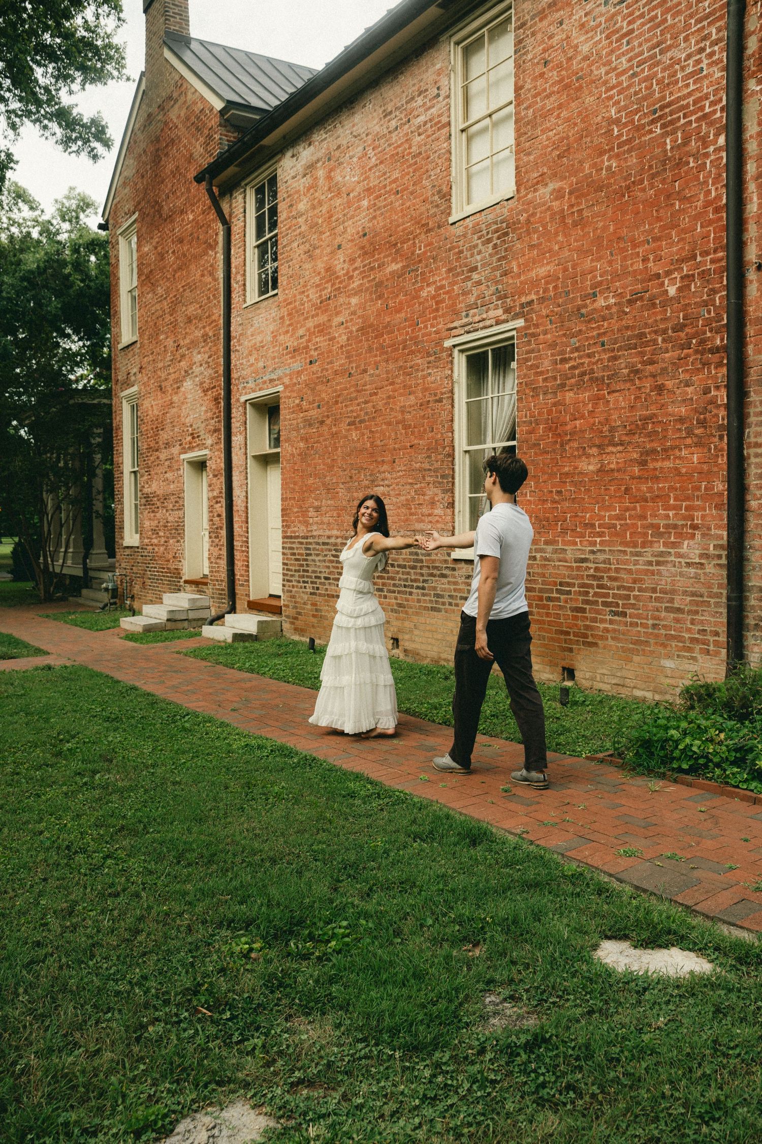 Couple dancing together on brick pathway beside historic red brick building with green lawn.
