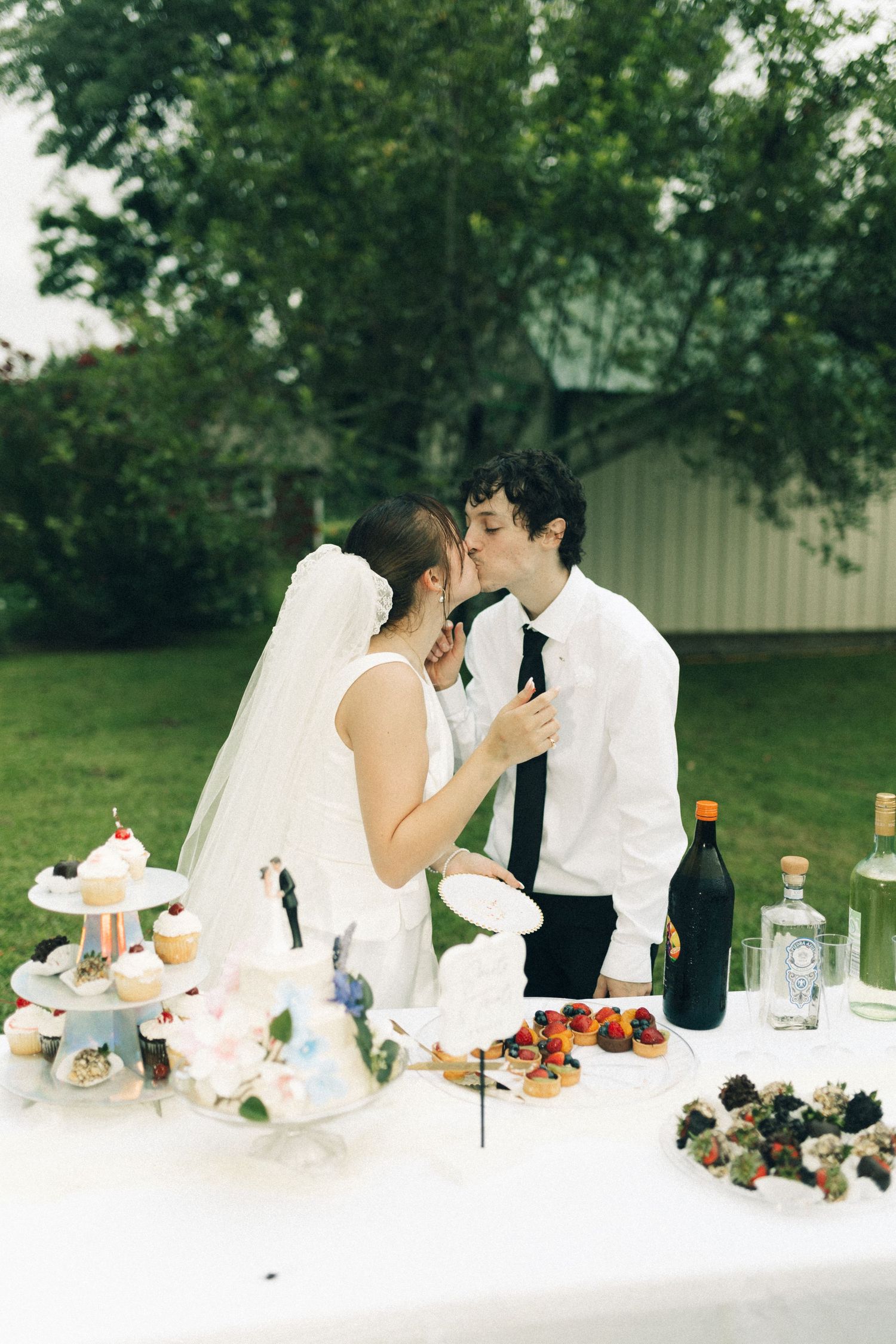 Newlyweds kissing at dessert table with cupcakes and treats at outdoor wedding reception.