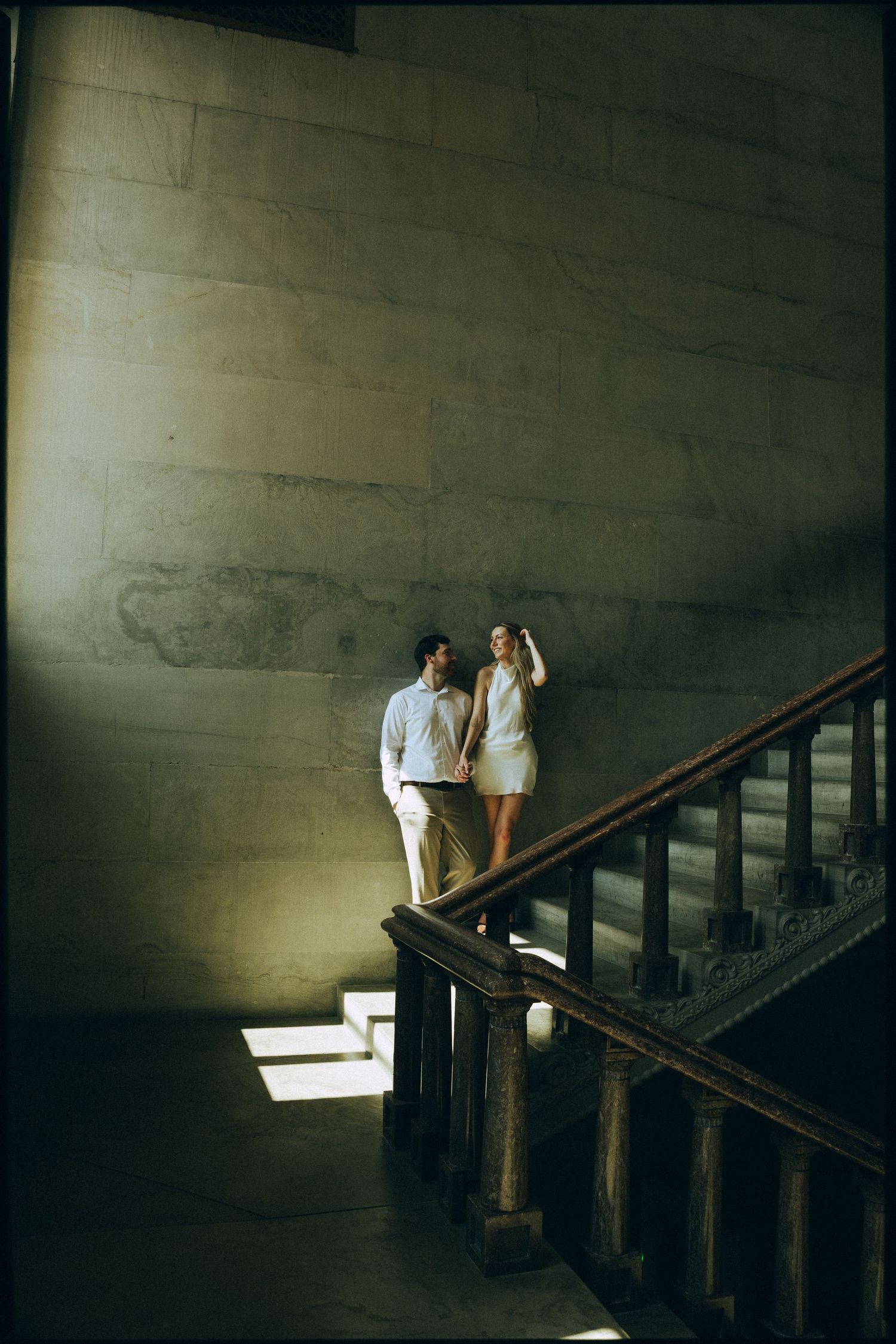 Couple holds hands on grand staircase bathed in dramatic light inside a stone building.