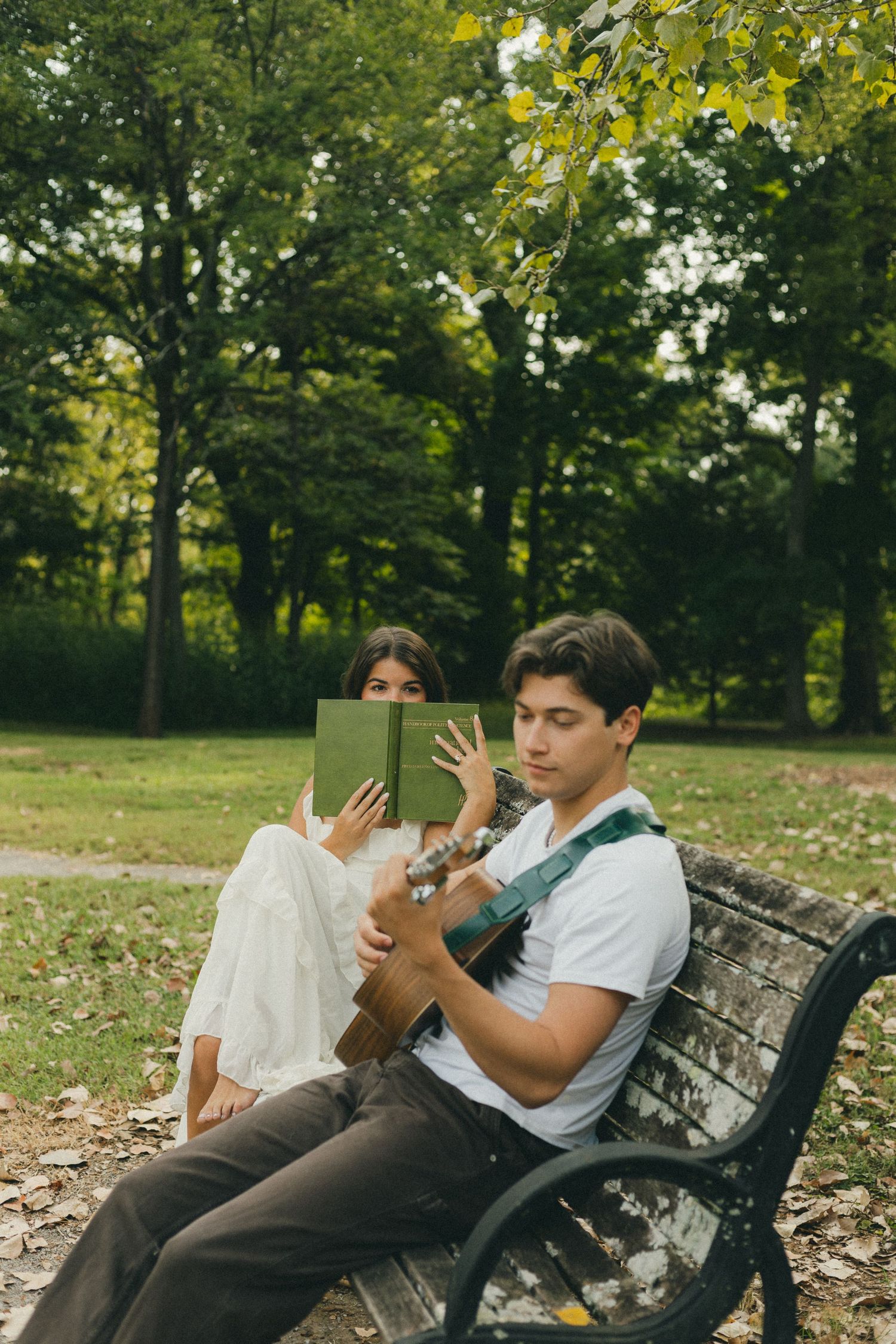 A young couple sits on a park bench, man playing guitar while woman reads a green book outdoors.