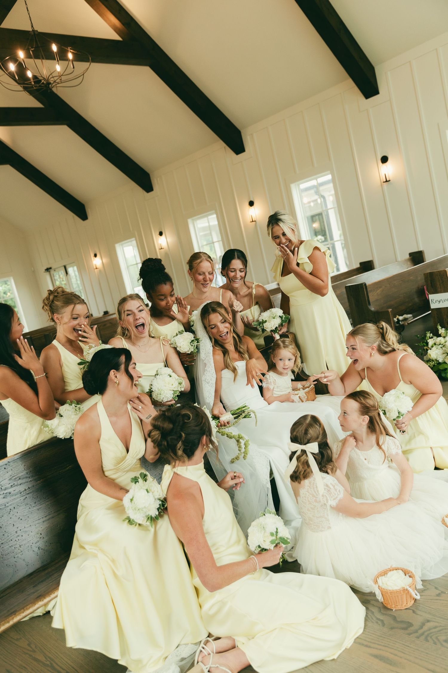 Joyful bride surrounded by bridesmaids in yellow dresses inside a rustic white chapel with exposed wooden beam ceiling.
