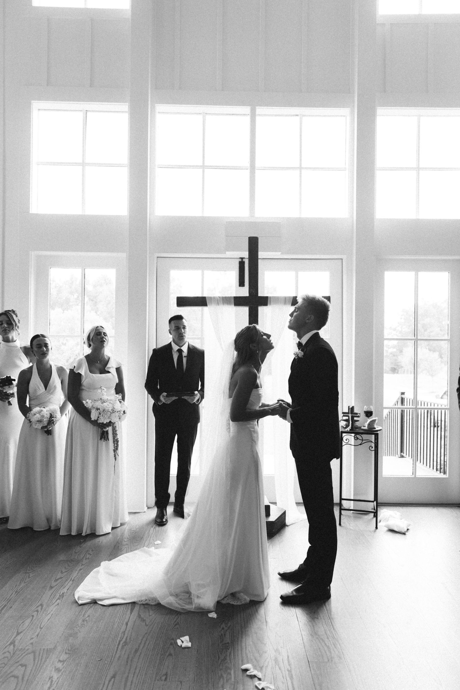 Bride and groom exchange vows before wooden cross in bright white chapel surrounded by wedding party.