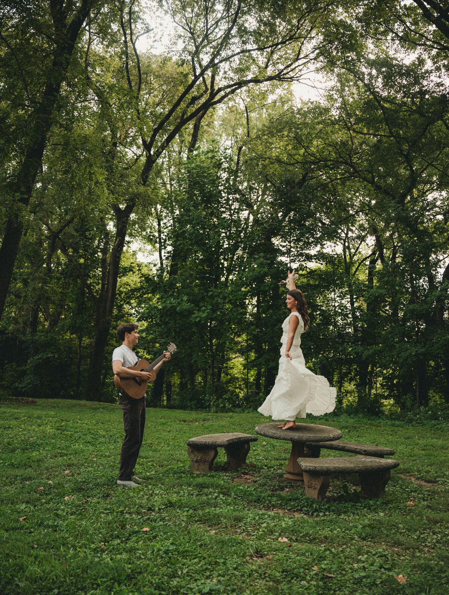 A groom plays guitar while a bride in a white dress dances on stone benches in a lush green park setting.