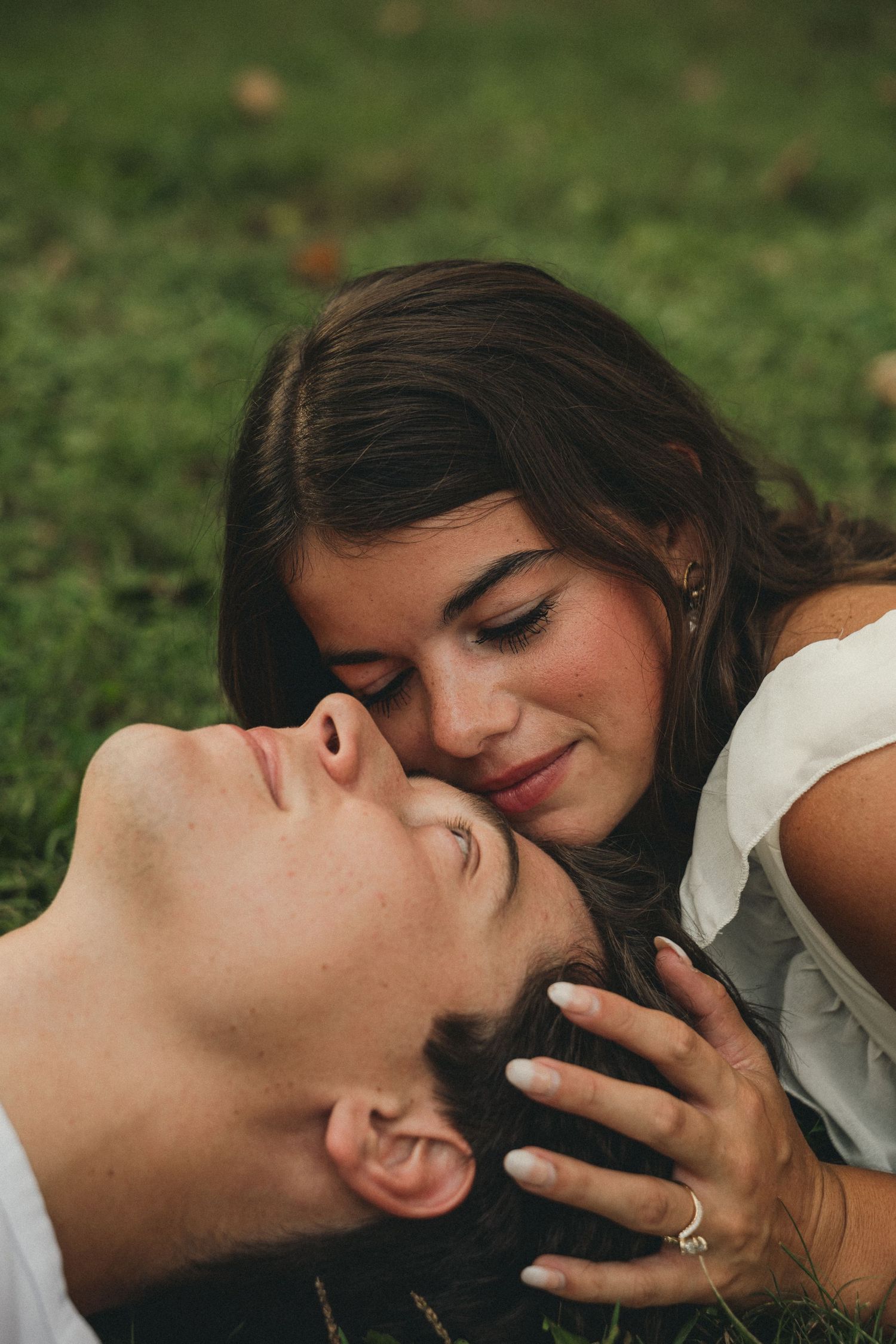 Romantic couple lying on grass, woman tenderly cradling mans head in intimate engagement photo.
