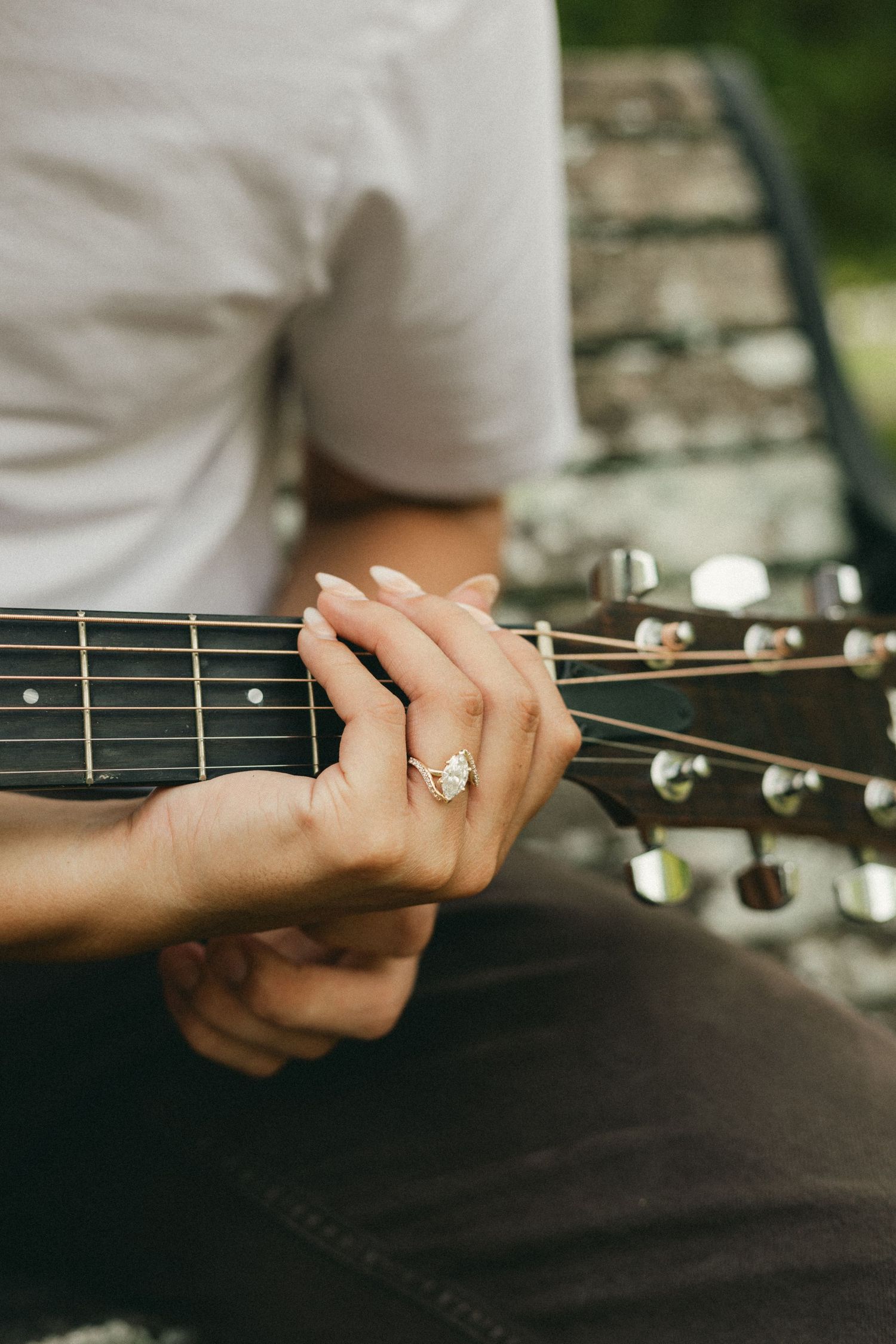 Close-up of a woman's hand with an engagement ring playing acoustic guitar strings outdoors.