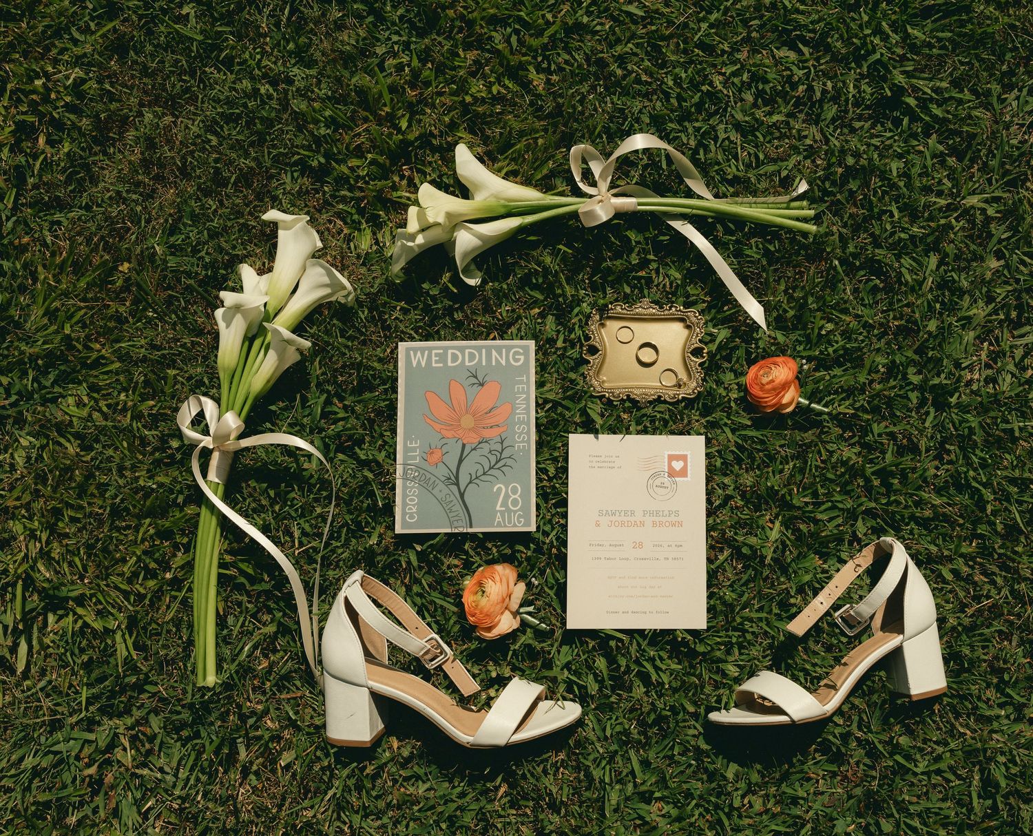Wedding flat lay on grass featuring white heels, calla lilies, rings, boutonniere, invitation suite, and vintage stamp card.
