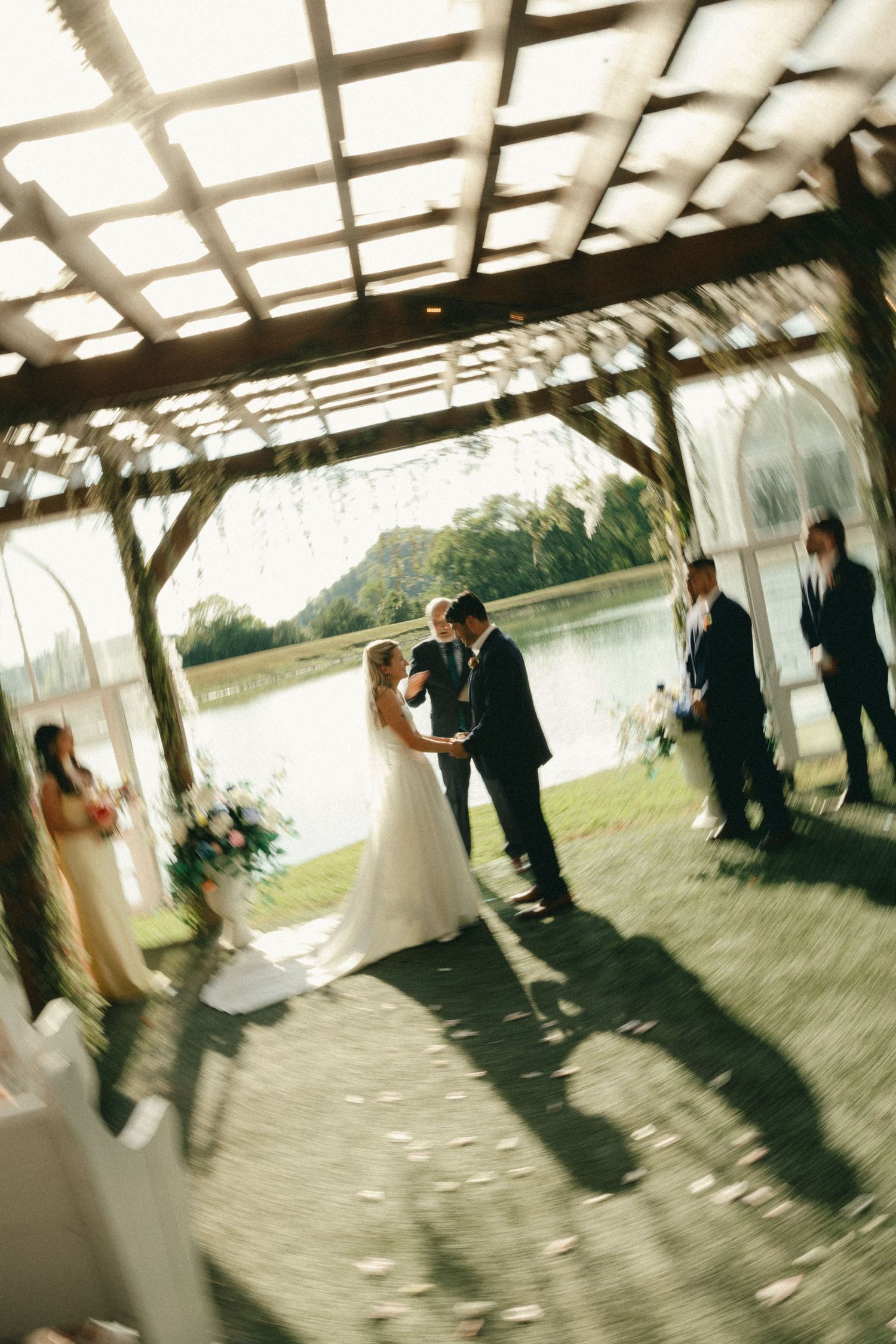 Bride and groom share first kiss under rustic pergola by lakeside during outdoor wedding ceremony.