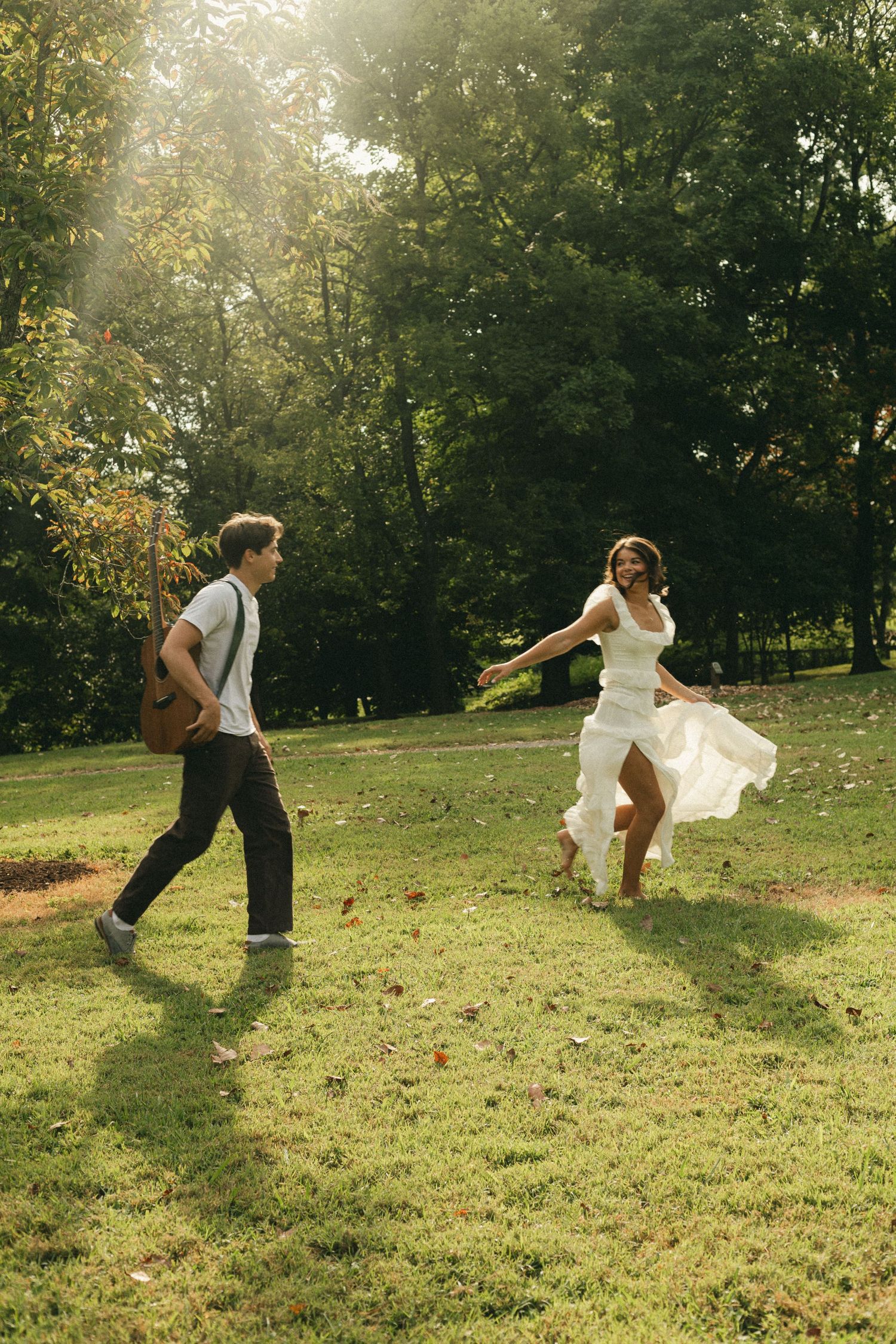 A joyful couple runs through a sunlit park, the woman in a flowing white dress and the man carrying a guitar.