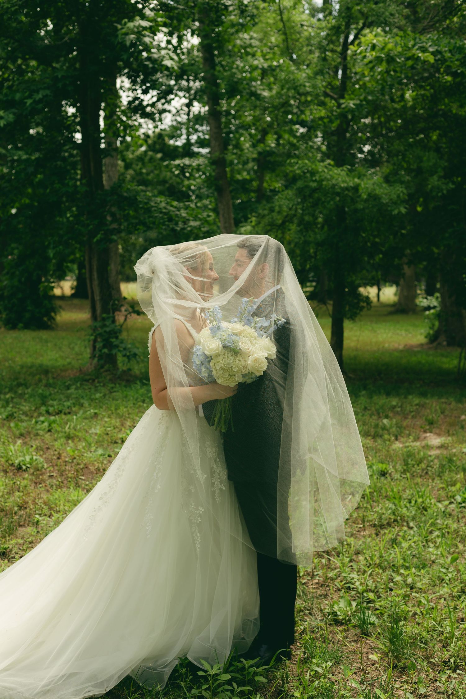 Bride and groom share a hidden kiss beneath the bride's flowing veil in a lush green wooded outdoor setting.