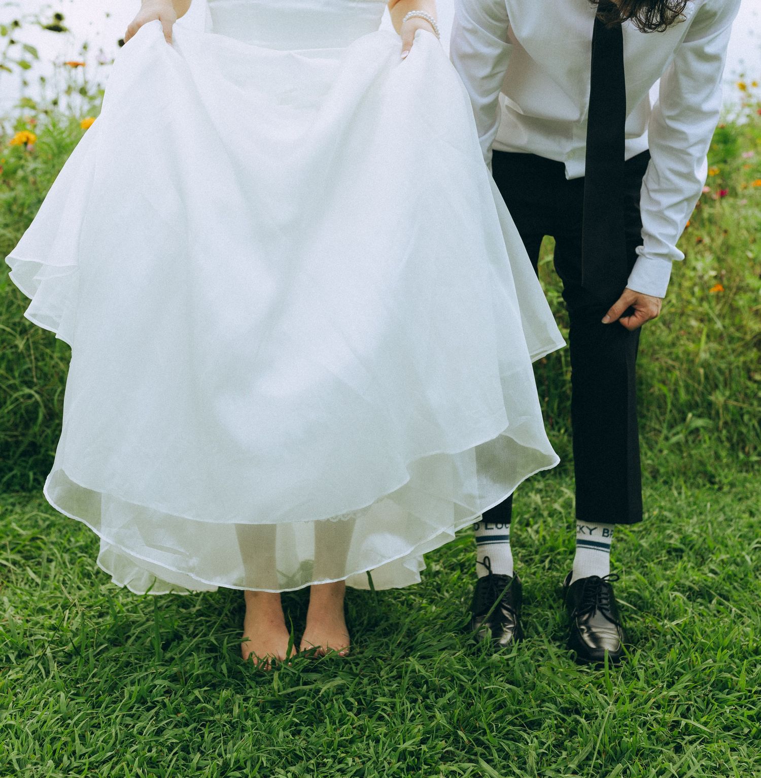 Bride in white gown lifts dress hem revealing bare feet beside groom in black pants and dress shoes on green grass.