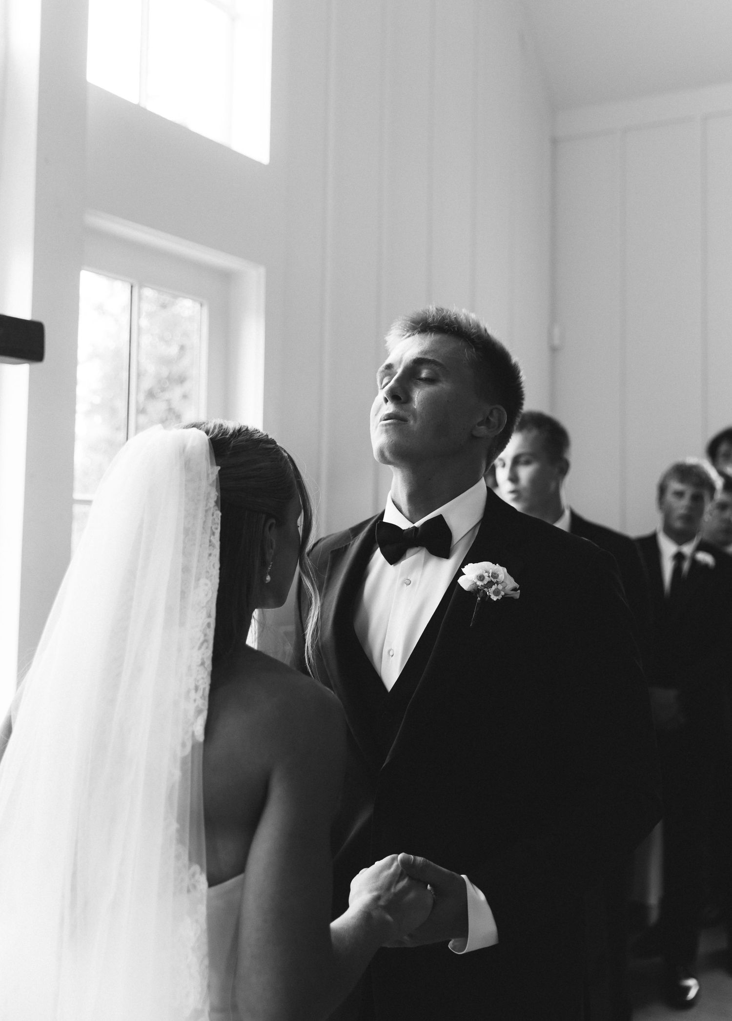 Emotional groom looks up during wedding vows while holding bride's hands at chapel altar in black and white.