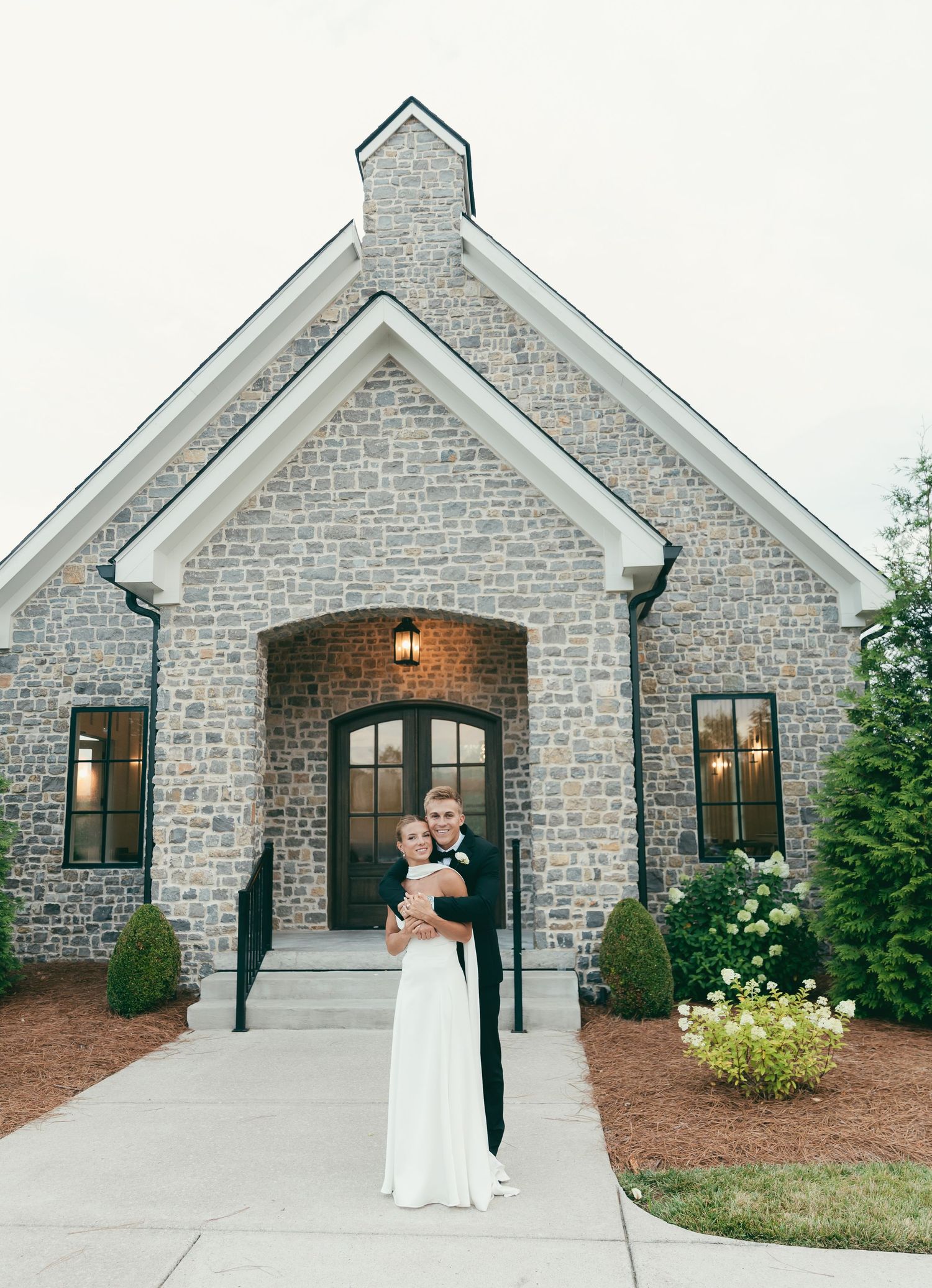 Bride and groom embrace in front of an elegant stone chapel with arched doors and manicured landscaping.
