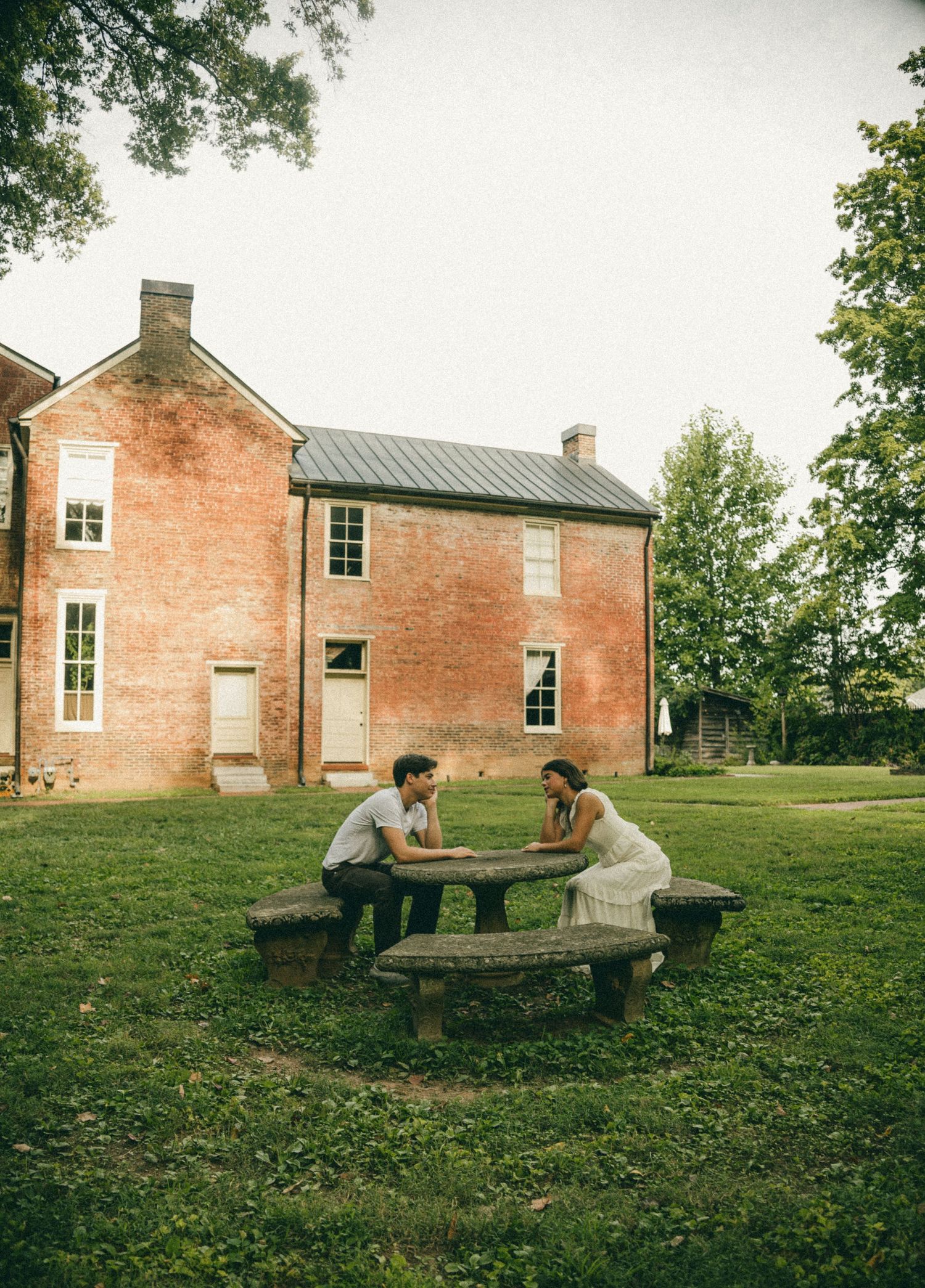 Couple sitting at a stone picnic table outside a historic red brick building surrounded by lush green trees and lawn.