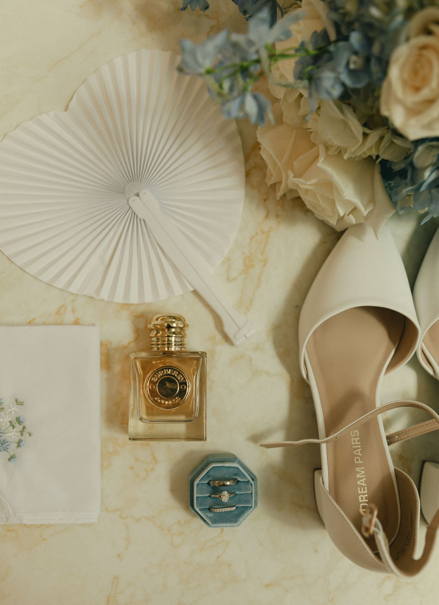 Wedding flat lay with perfume bottle, blue velvet ring box, white heels, fan, and floral bouquet.