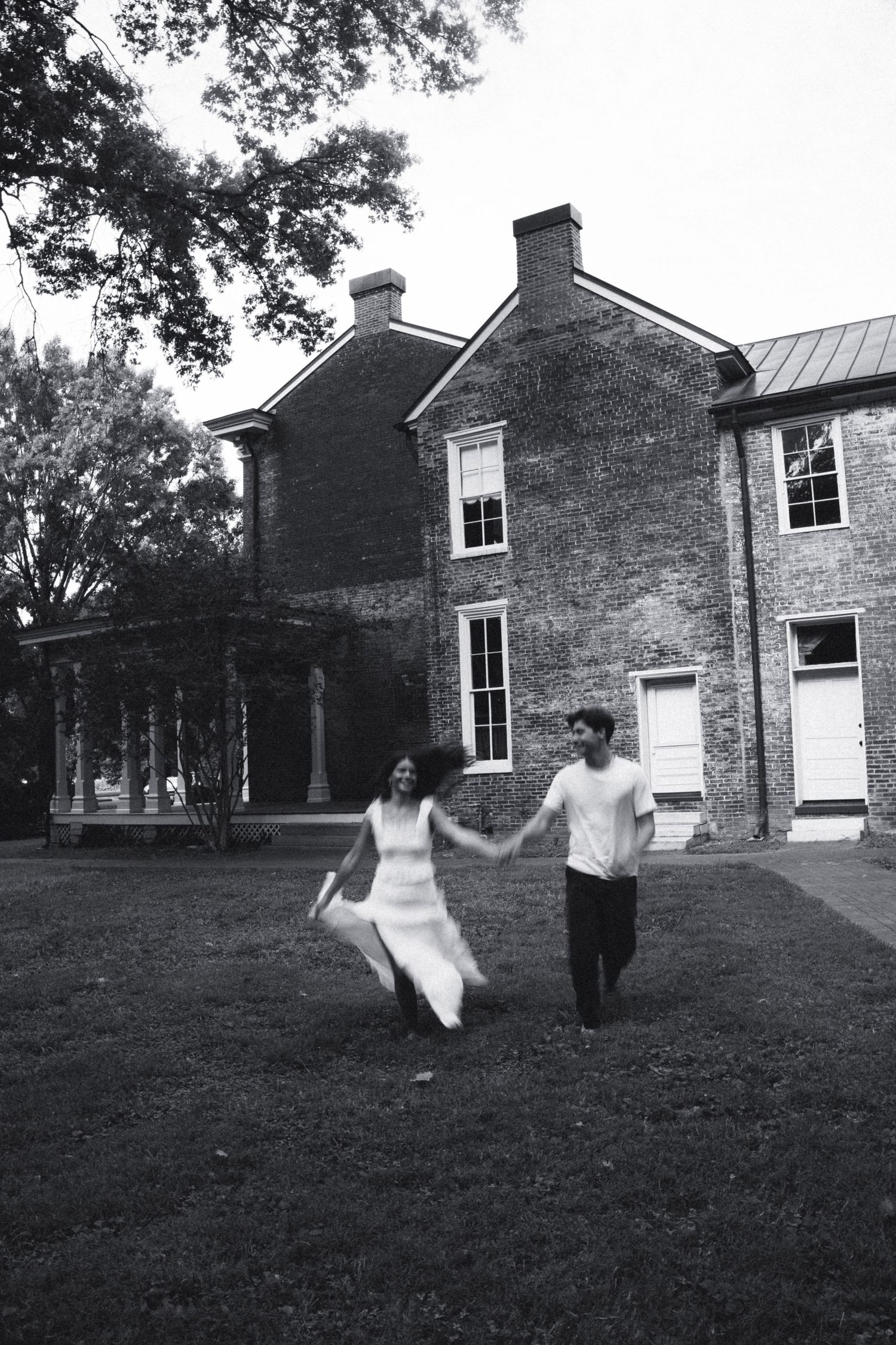 Black and white photo of couple holding hands running across lawn of historic brick mansion.