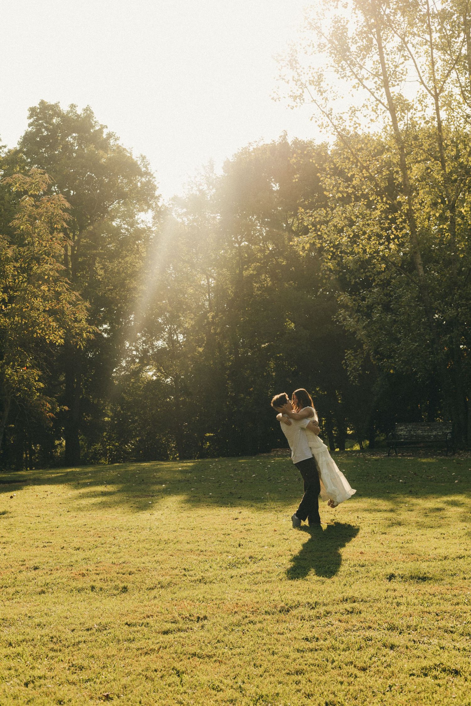 A couple shares a romantic embrace in a sunlit park, with golden light filtering through lush green trees.