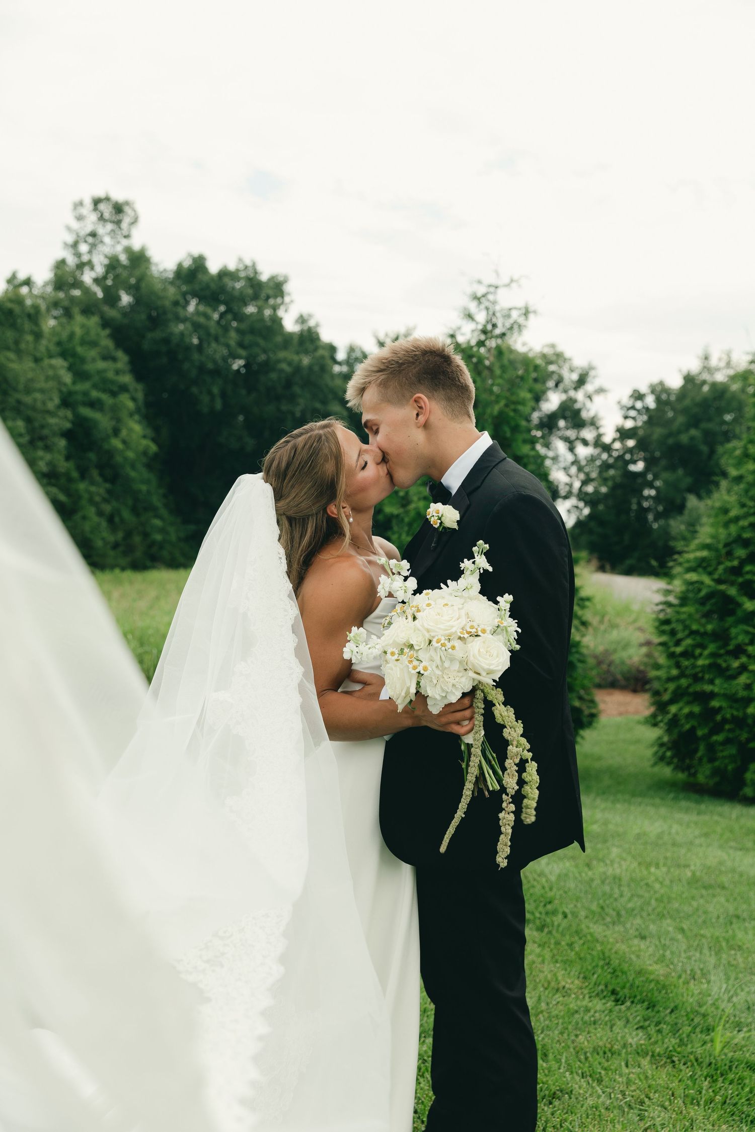 Bride and groom share a romantic kiss outdoors, bride holding a white floral bouquet with her lace veil flowing beside them.