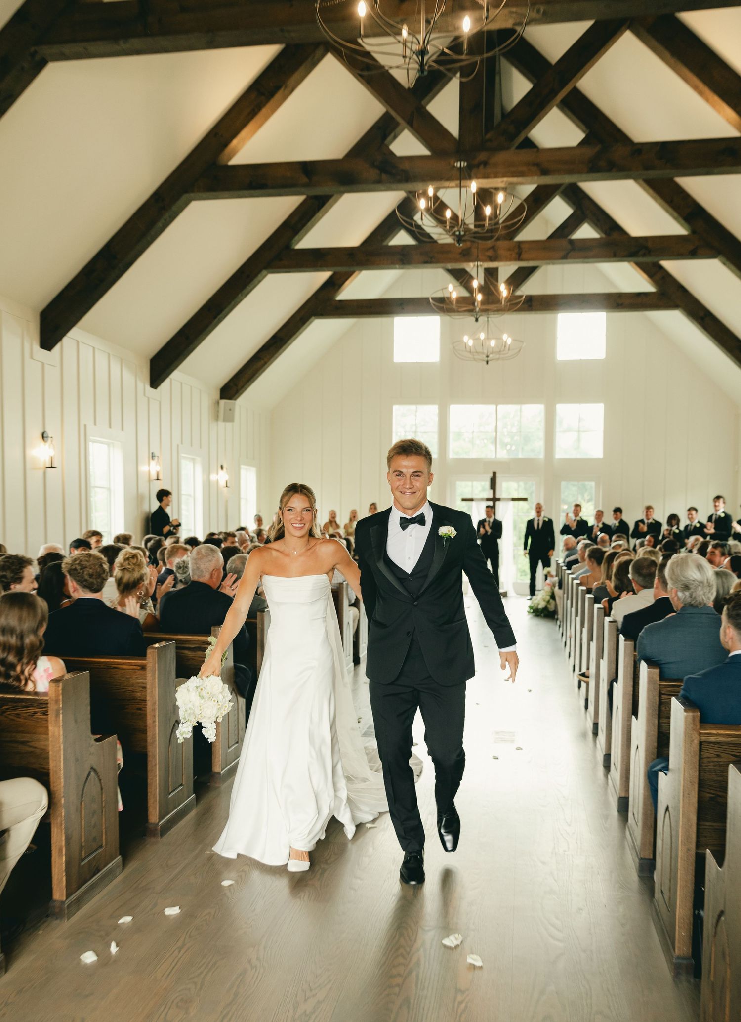 Newlyweds walk joyfully down aisle after ceremony in elegant white chapel with exposed wood beam ceiling.