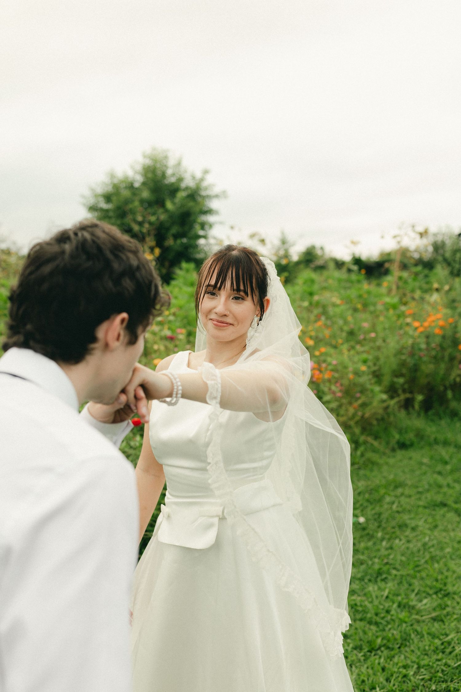 Groom kissing bride's hand in garden while she smiles wearing white wedding dress and veil.