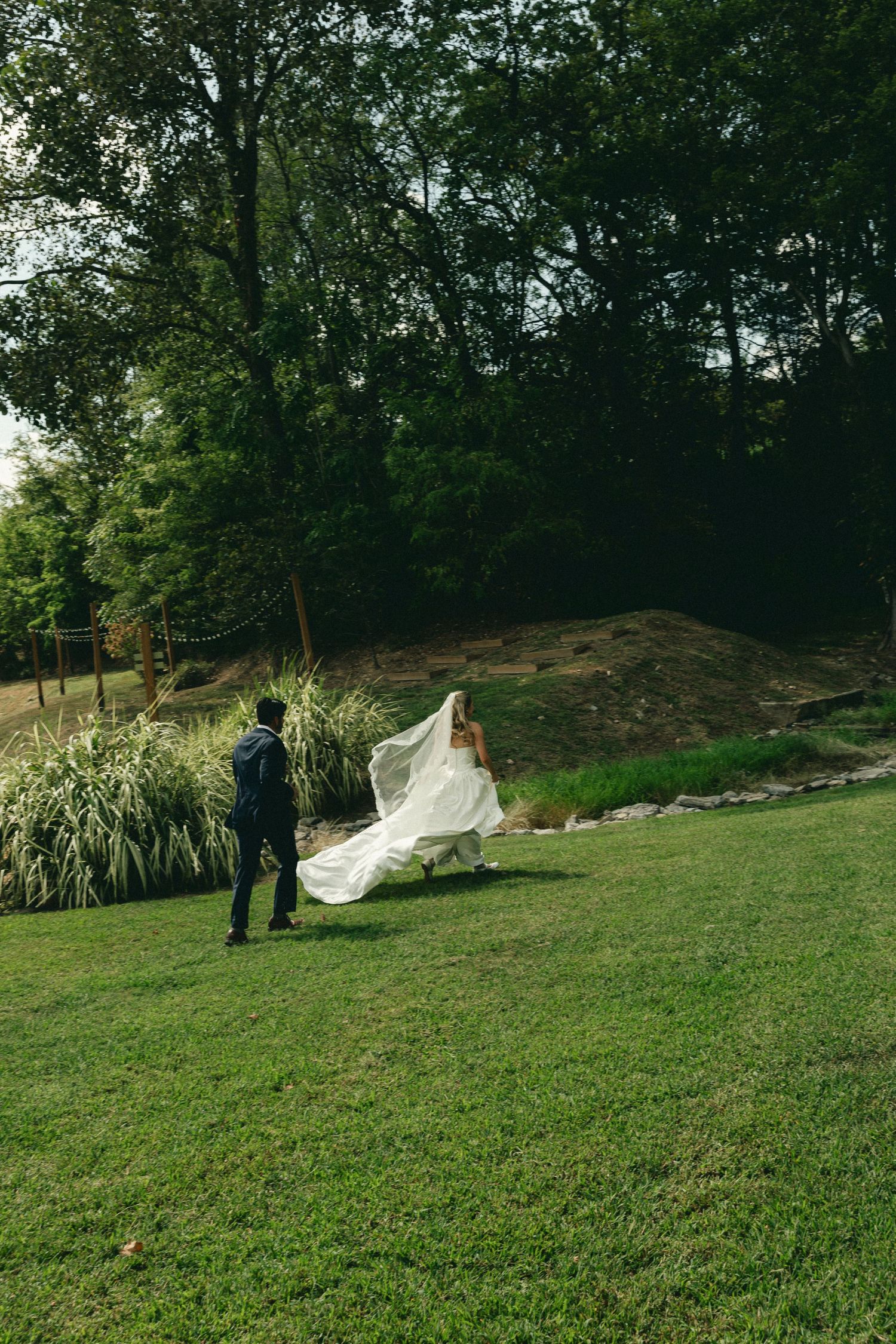 Bride in flowing white gown and veil walks with groom across lush green lawn surrounded by trees.
