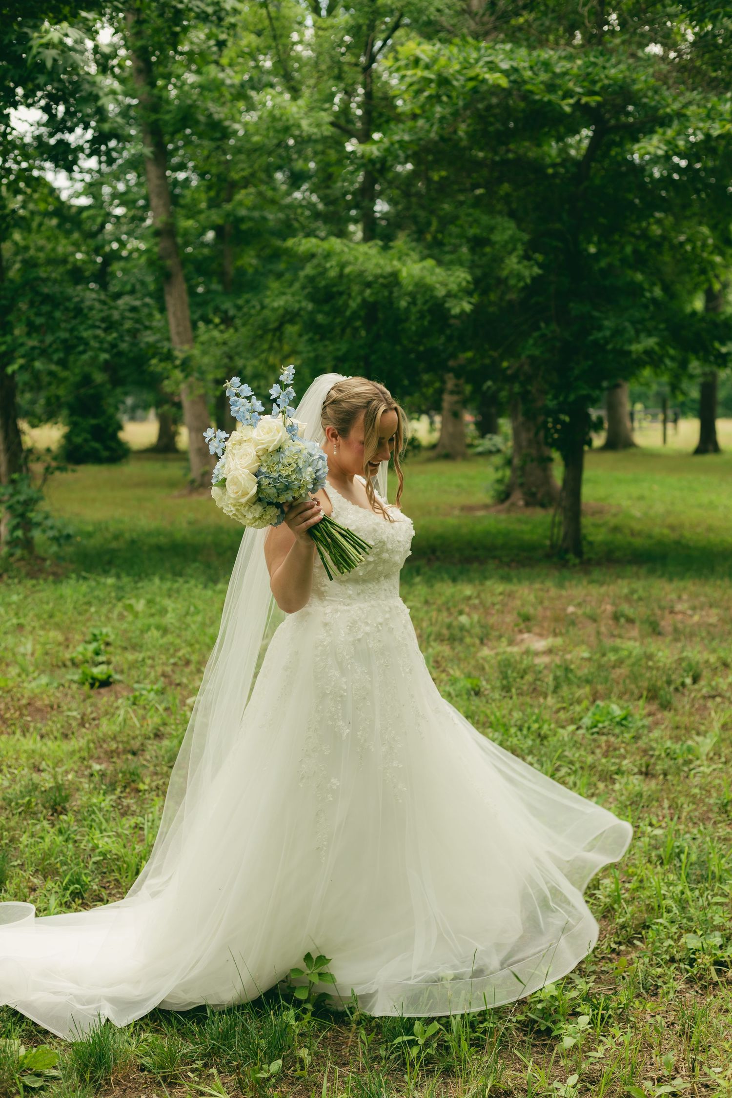 Elegant bride in a lace ball gown holding a blue and white bouquet while standing in a green wooded outdoor setting.