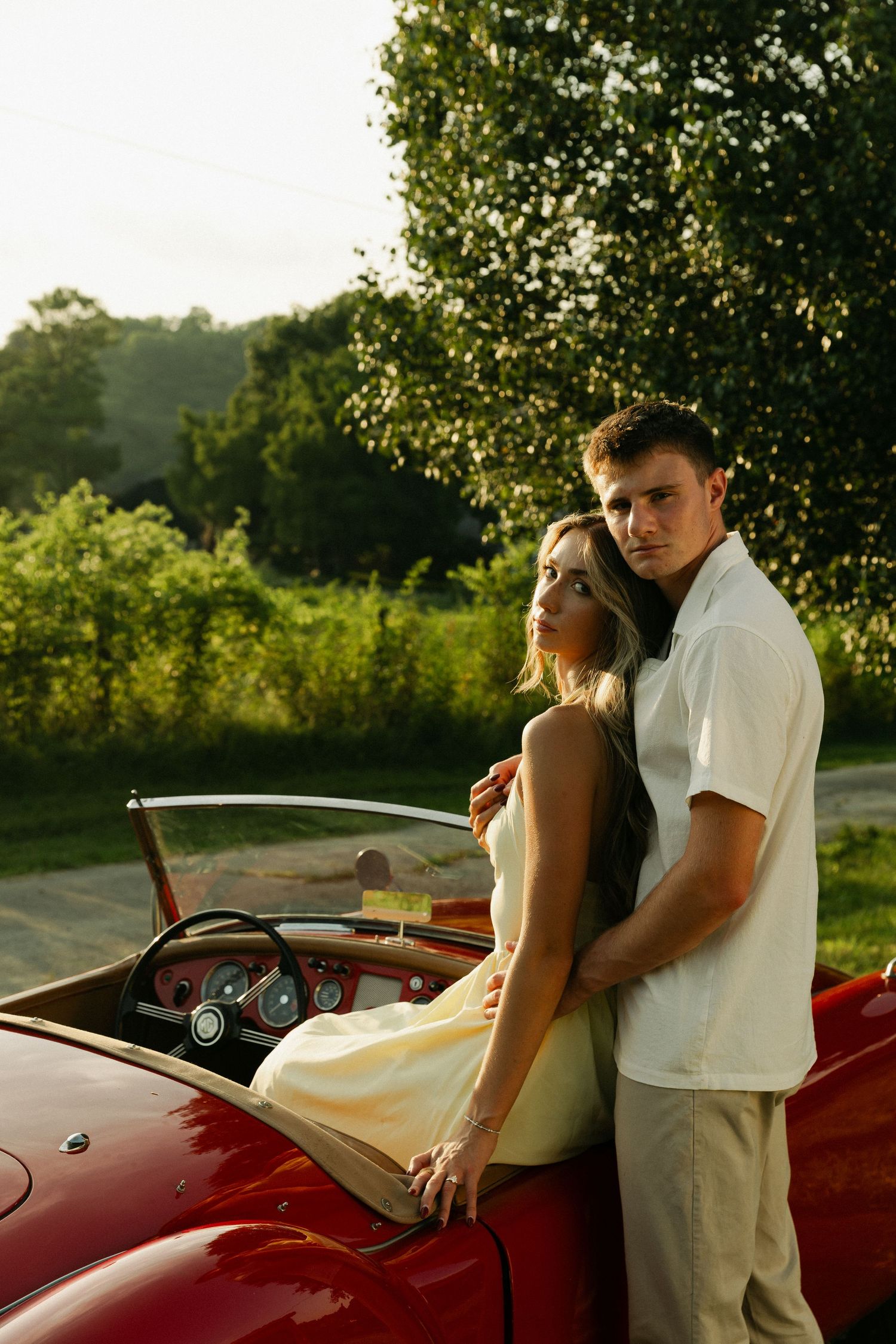 Romantic couple embracing beside a classic red vintage convertible sports car at golden hour.