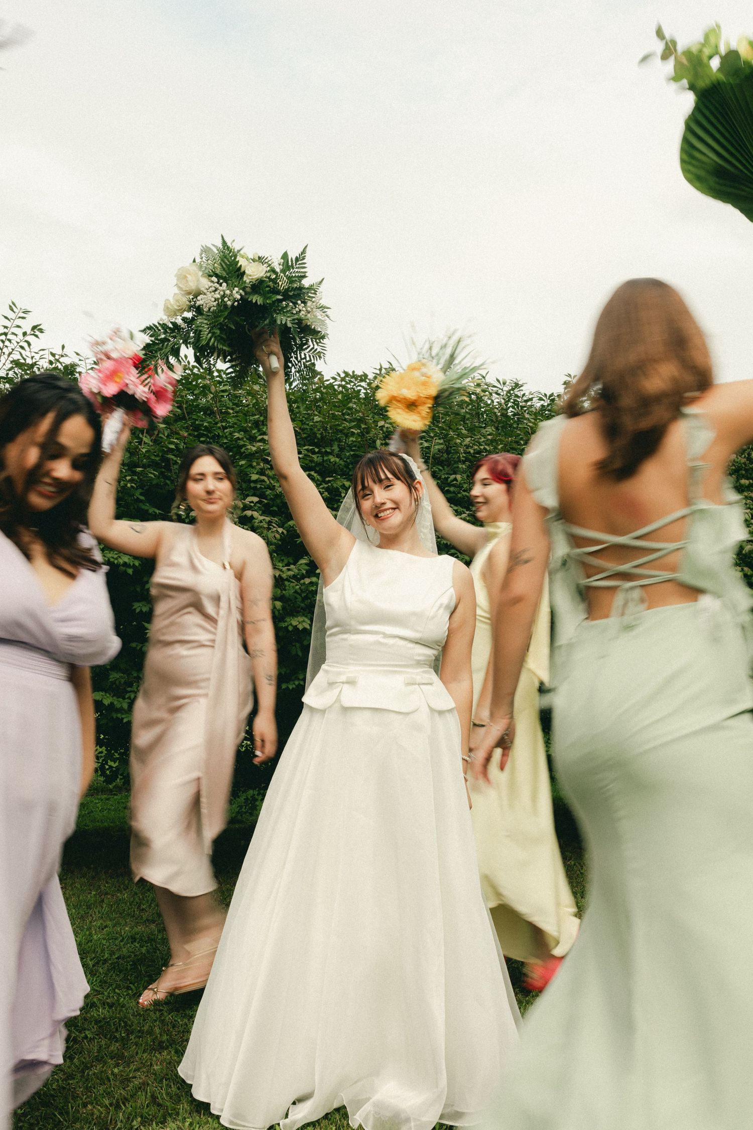 Joyful bride raising bouquet with bridesmaids celebrating outdoors at wedding reception.