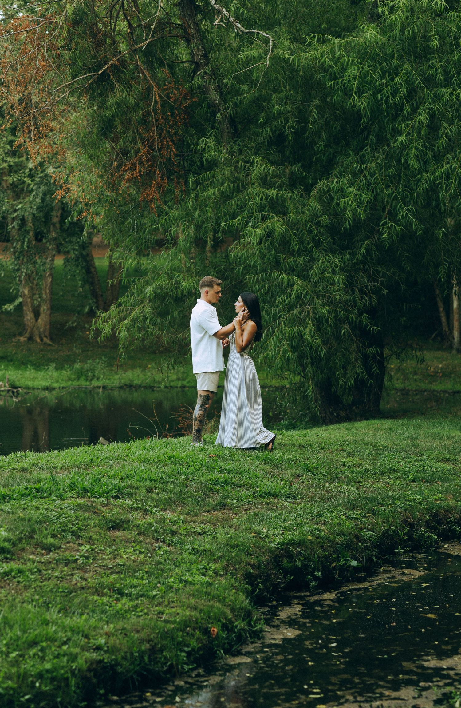 Couple embracing near pond surrounded by lush green weeping willow trees outdoors.
