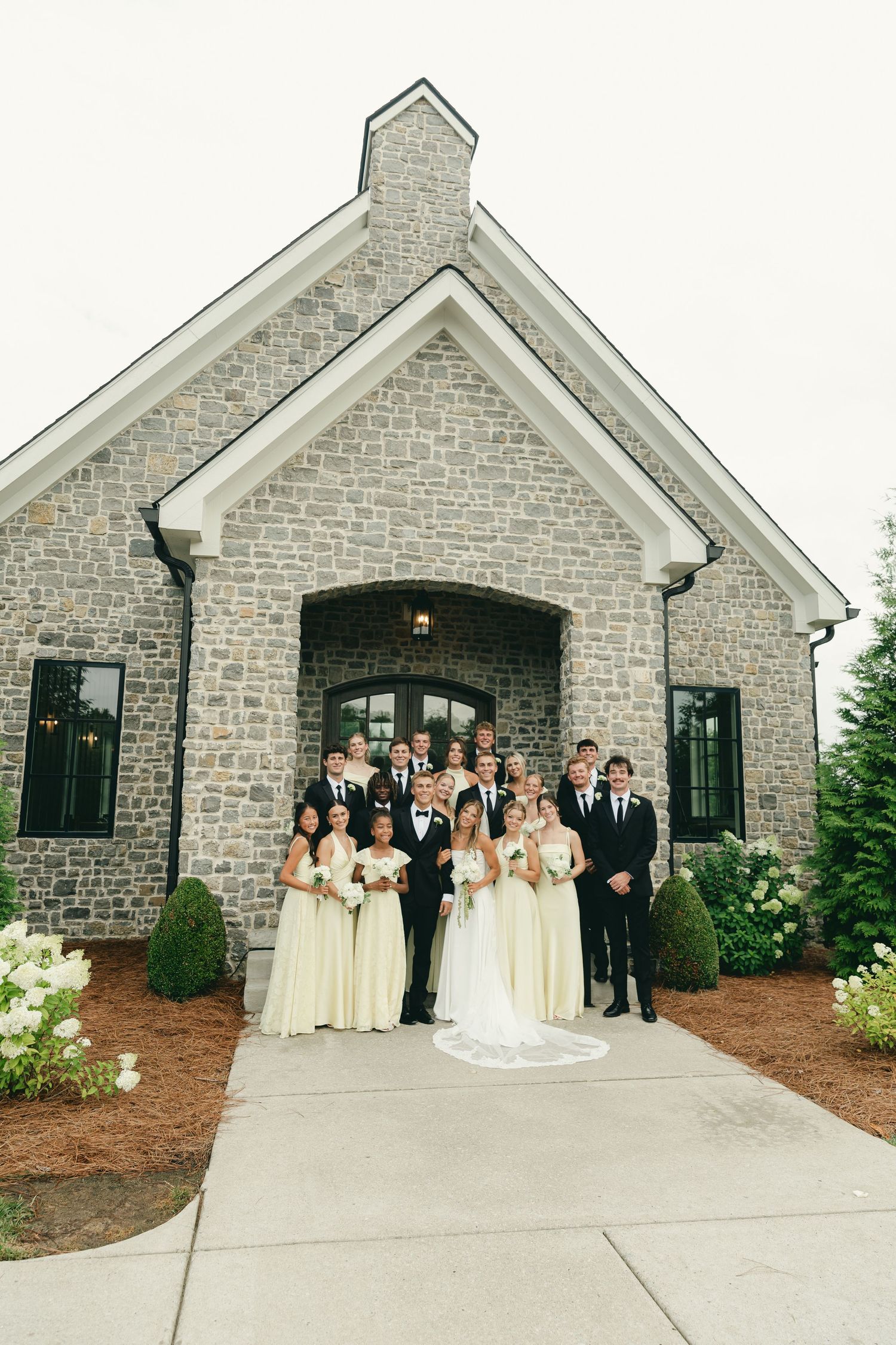 Full wedding party poses in front of a stone chapel on the couple's wedding day.