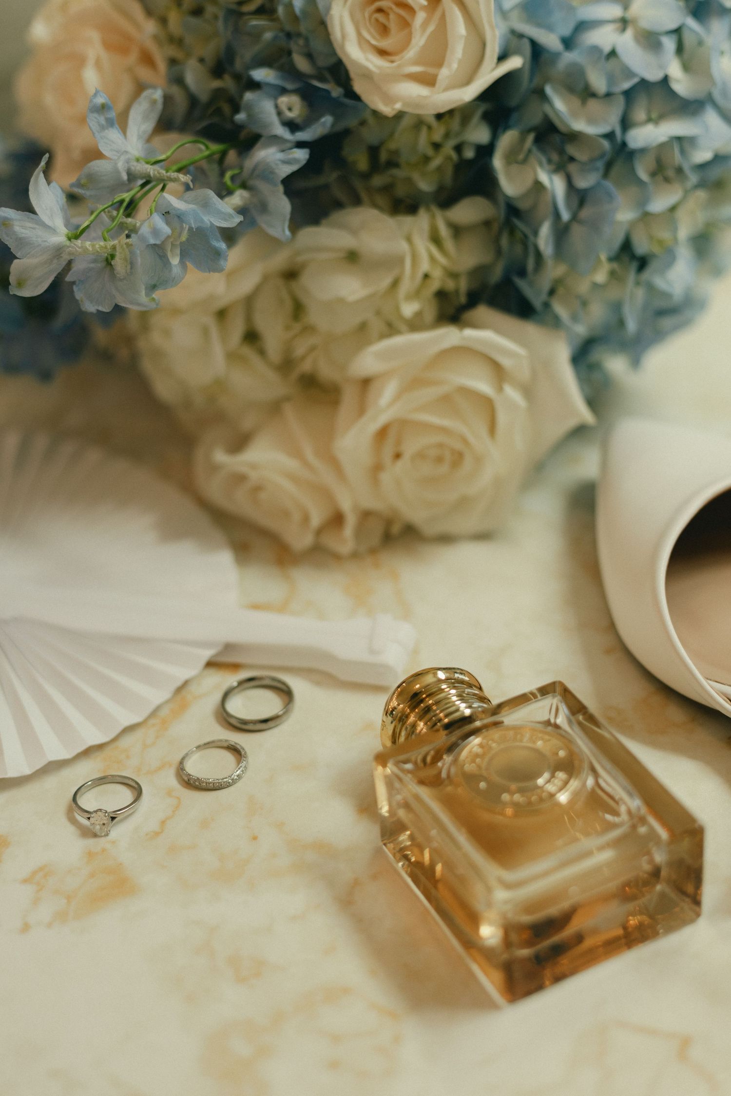 Wedding details flat lay featuring perfume bottle, rings, white roses, blue hydrangeas, and bridal heels.