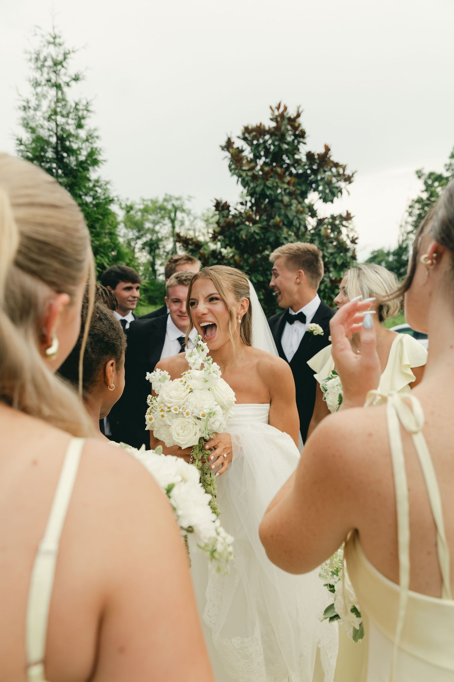 Joyful bride in white gown laughing with wedding party outdoors holding white floral bouquet.