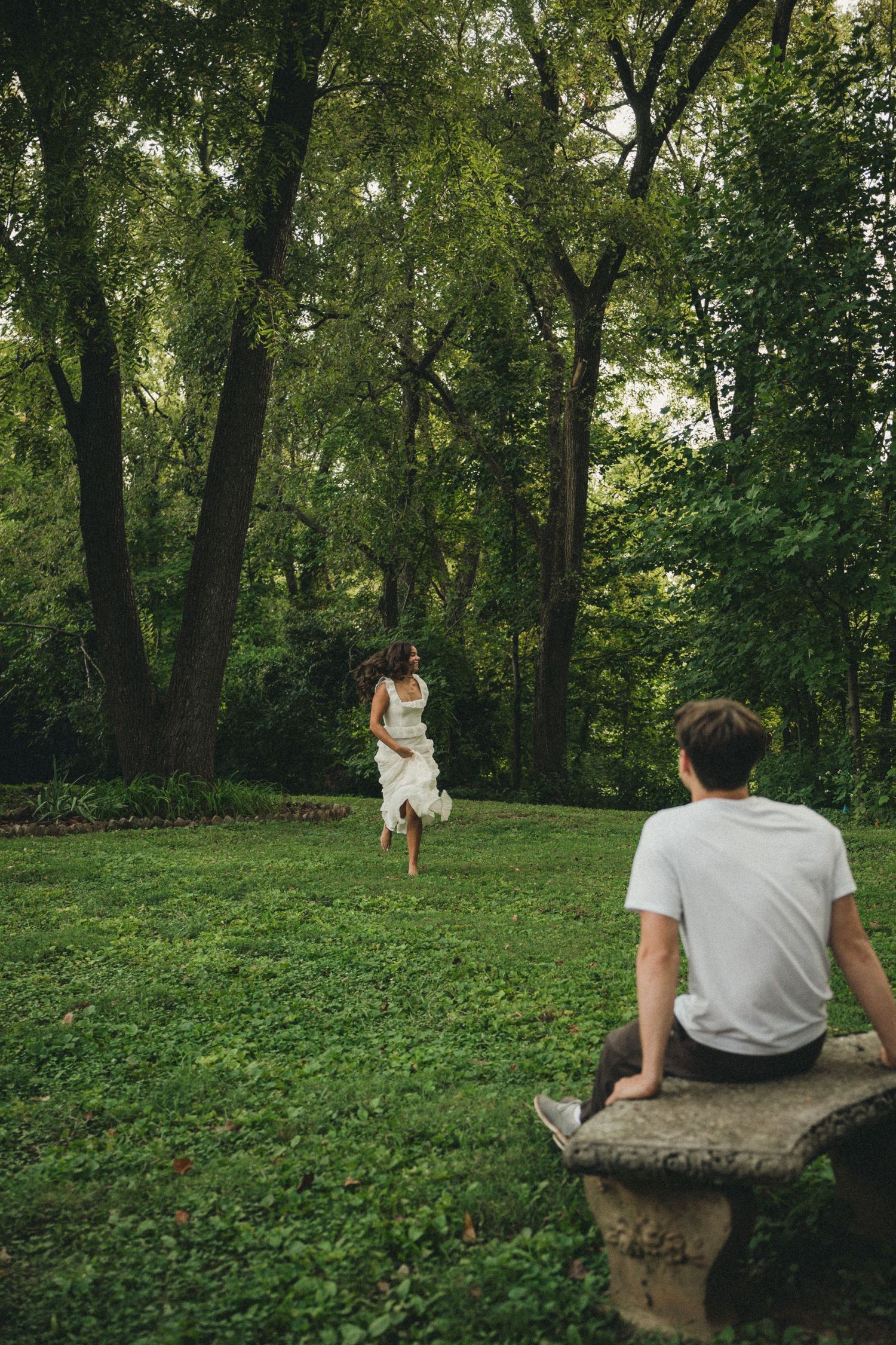 A woman in a white dress runs toward a man sitting on a stone bench in a lush green park.